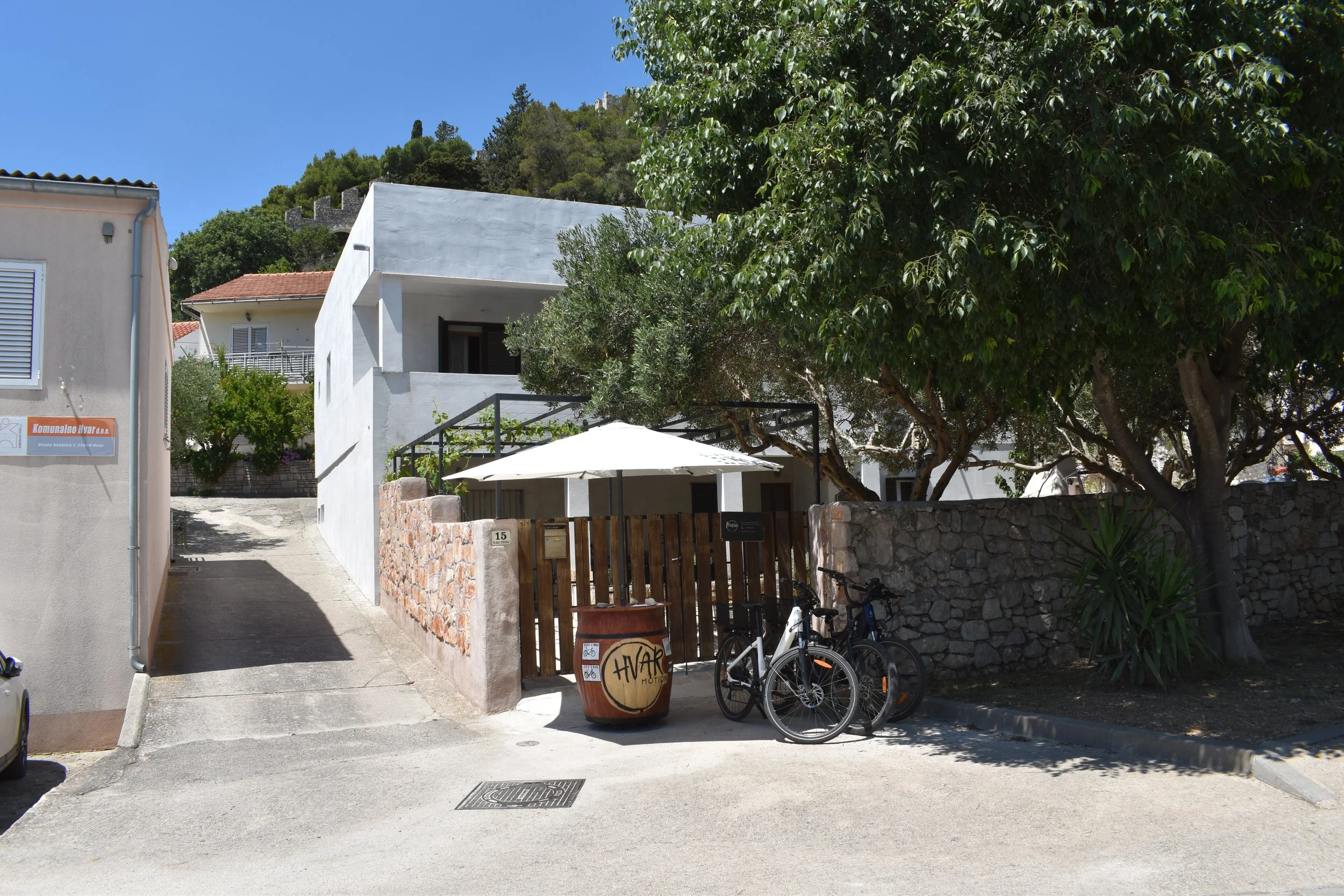 A narrow street with a residential white house on the right, a bicycle and a sign on a barrel in front, lush green trees on the right, and a hillside with greenery and ruins in the background under a clear blue sky.