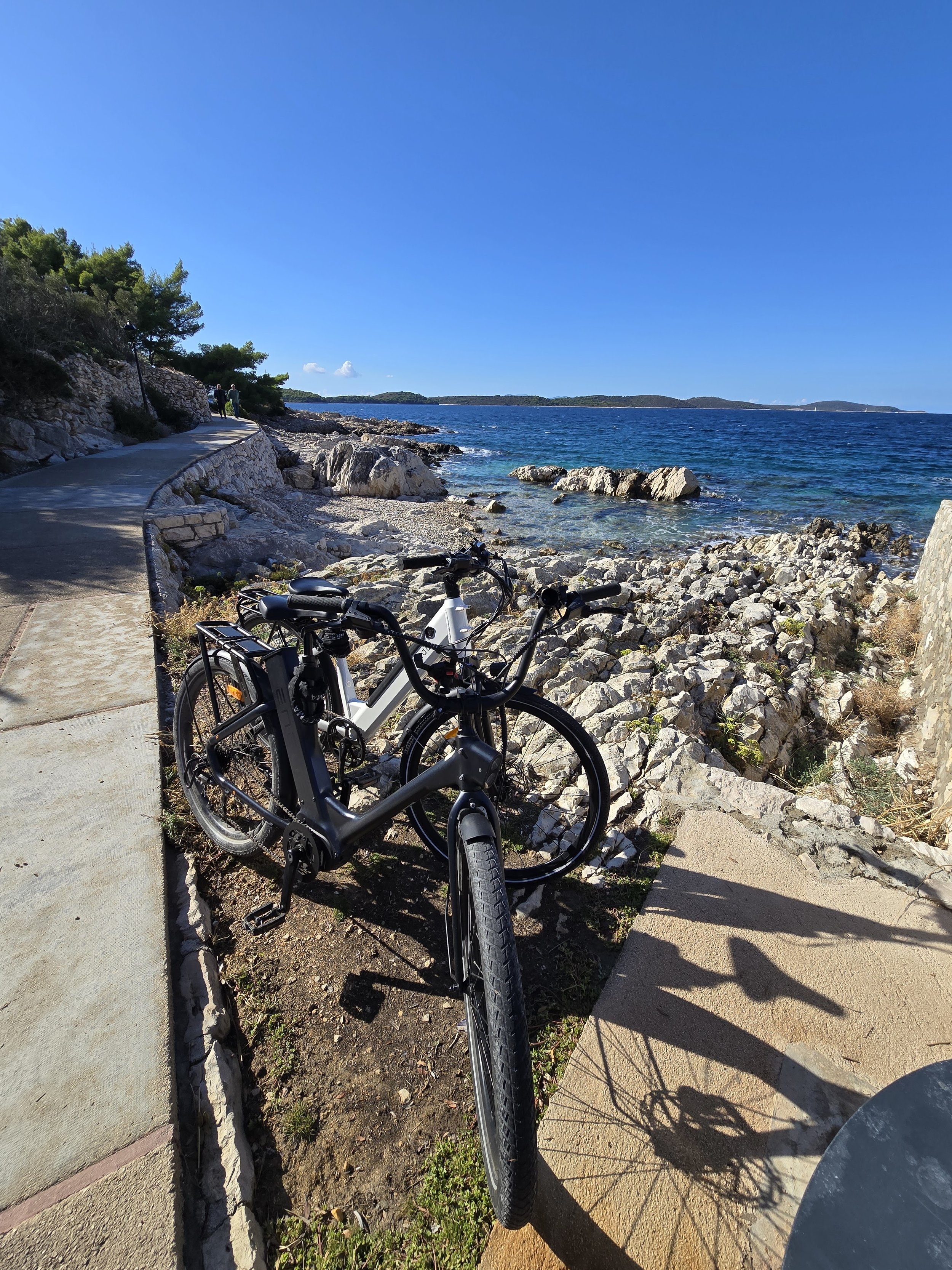 Bicycle parked on a rocky and sandy shoreline next to a paved walkway, overlooking a blue ocean with small islands in the distance under a clear blue sky.