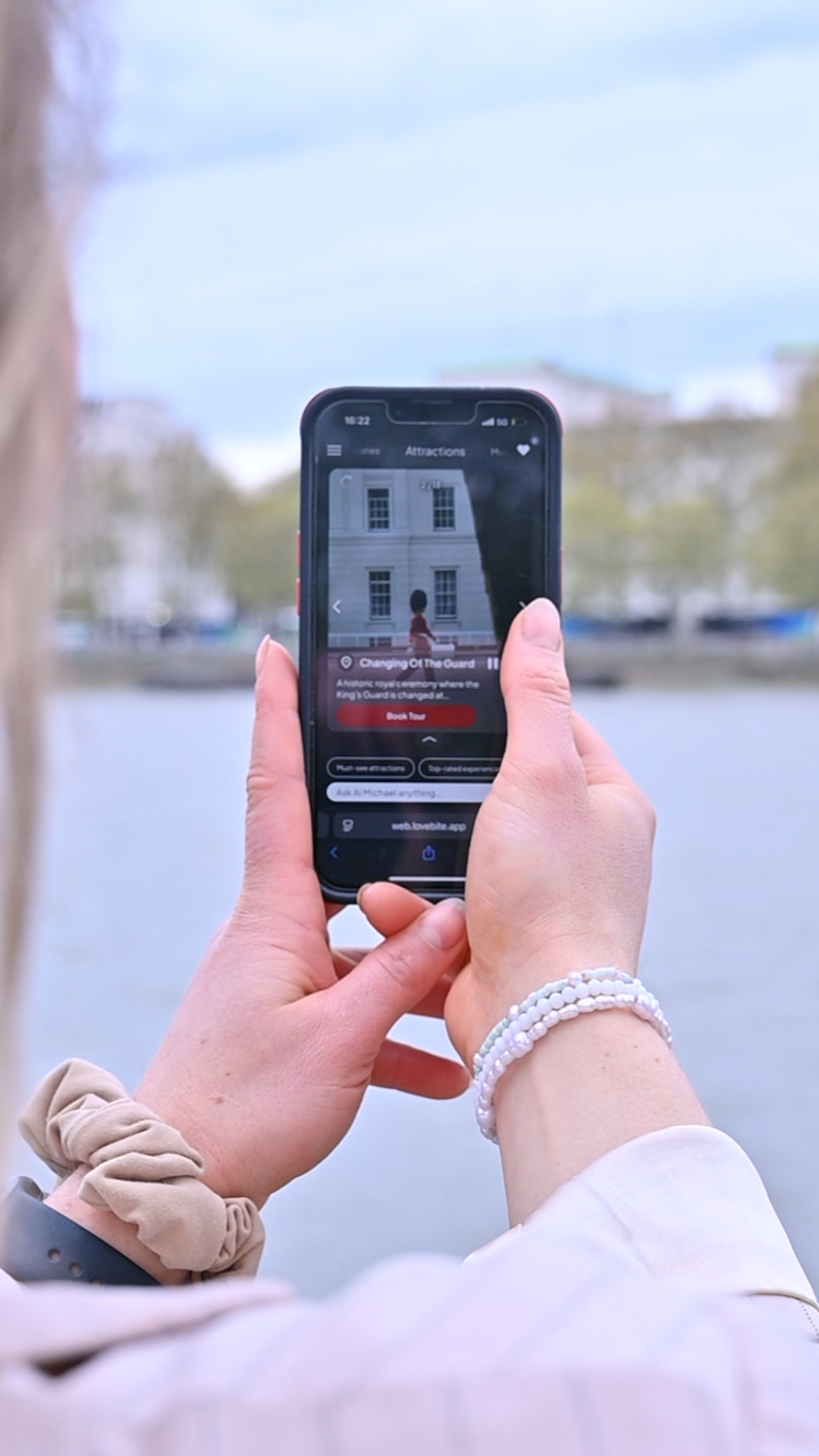 A visitor using a smartphone near the River Thames with a landmark in the background.