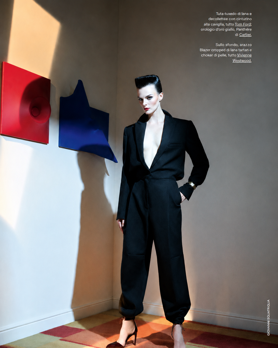 Fashion model in a black suit posing beside red and blue sculptural wall artworks in a contemporary interior with dramatic geometric lighting.
