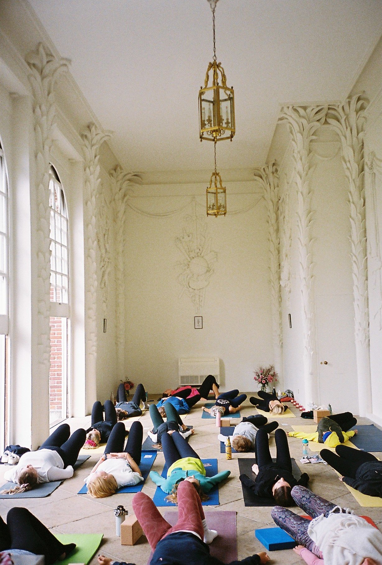 People participating in a yoga class in a spacious, decorated room with tall windows and ornate white walls. Participants are lying on mats in a relaxed pose.