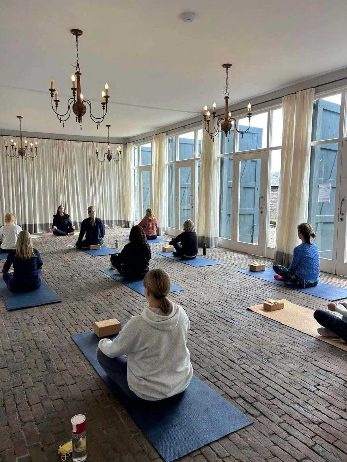 People participating in a yoga or meditation class in a spacious room with large windows, chandeliers, brick floor, and cream curtains.
