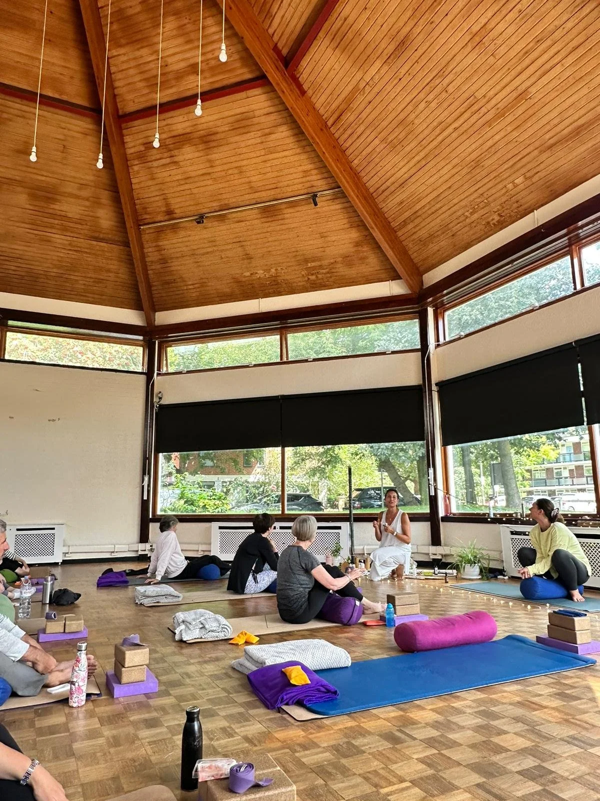 People participating in a yoga class in a spacious room with wooden ceiling, large windows, and mats on the floor.