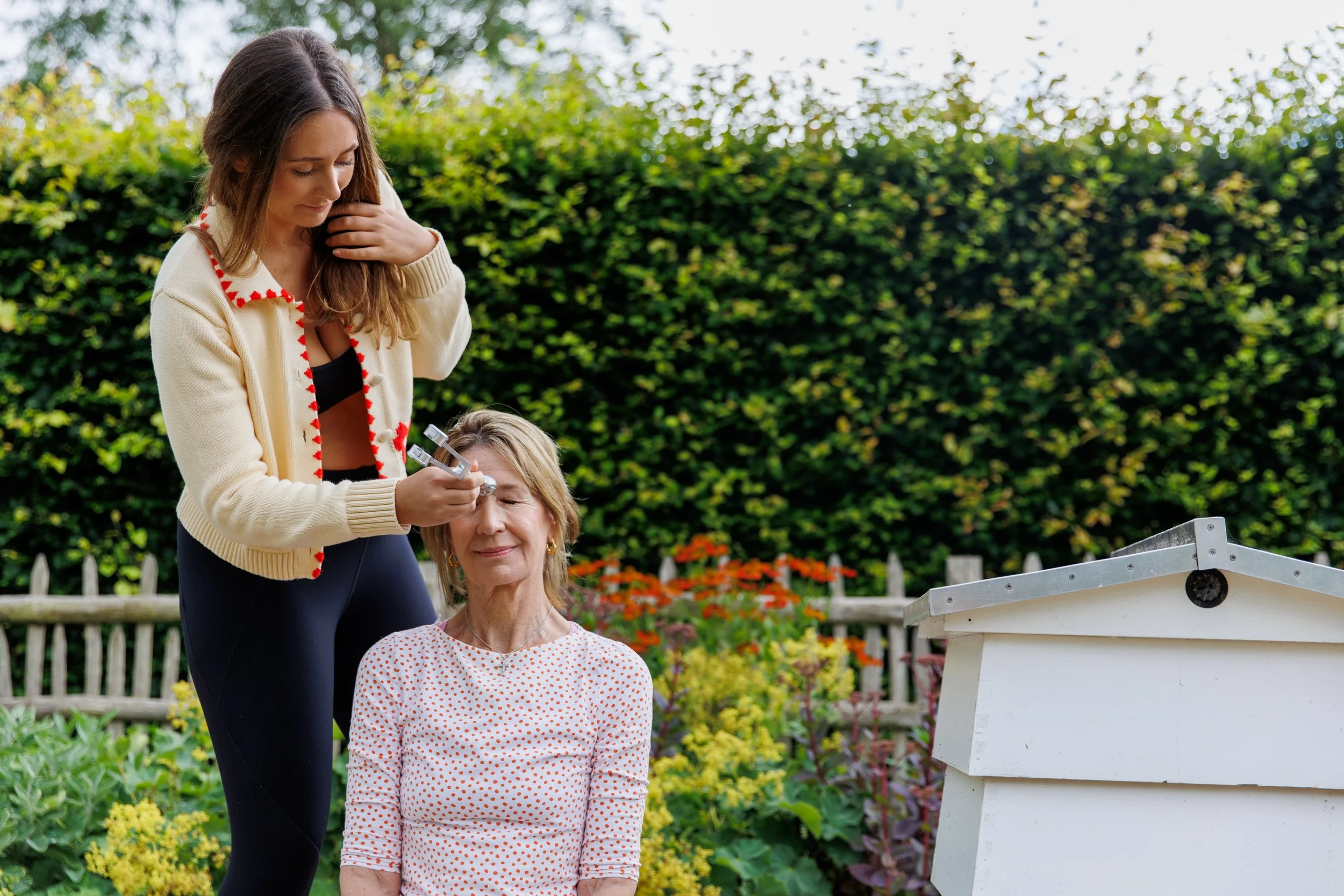A woman in a blue top and orange crochet cover-up stands in a garden full of purple and white flowers, with a wooden fence and a brick house with a tiled roof in the background, reaching upward with her right arm and eyes closed.