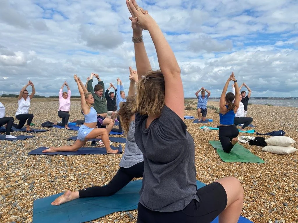 Group of people practicing yoga on a pebbled beach under cloudy sky, with some in lunge poses and others with hands raised in up position.