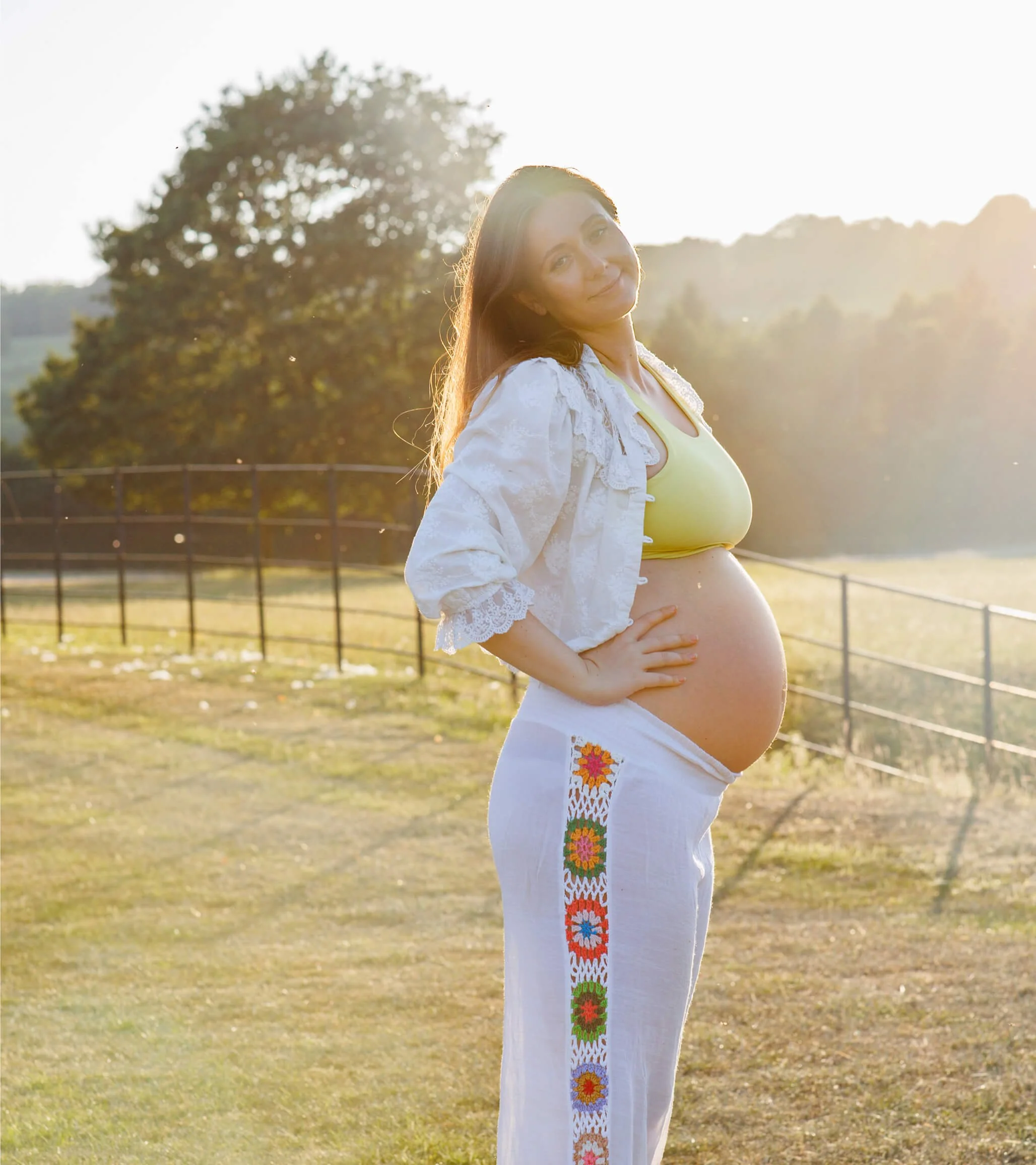 Pregnant woman standing outdoors in a field with a fence and trees in the background, sunlight shining behind her, wearing a yellow top and white embroidered pants.
