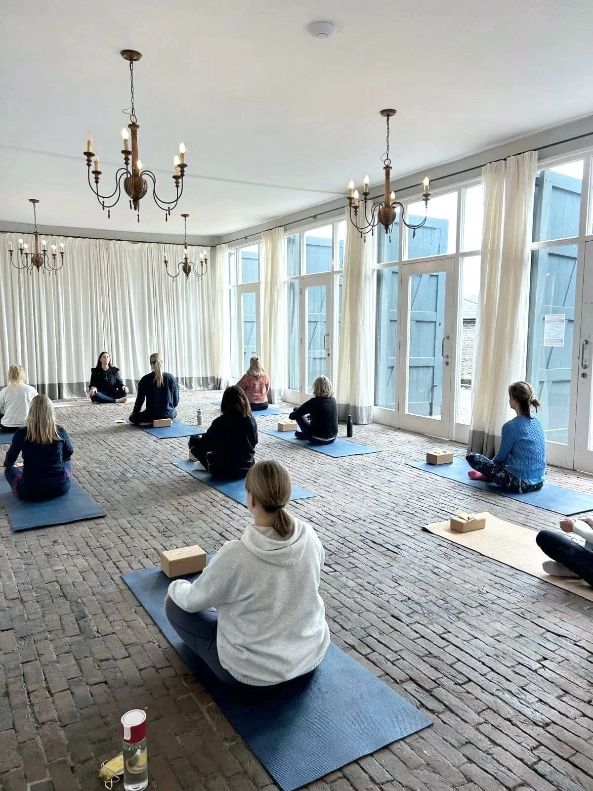 A group of people participate in a yoga or mindfulness class in a bright, spacious room with ornate white walls and large windows, overseen by a woman in pink guiding them.