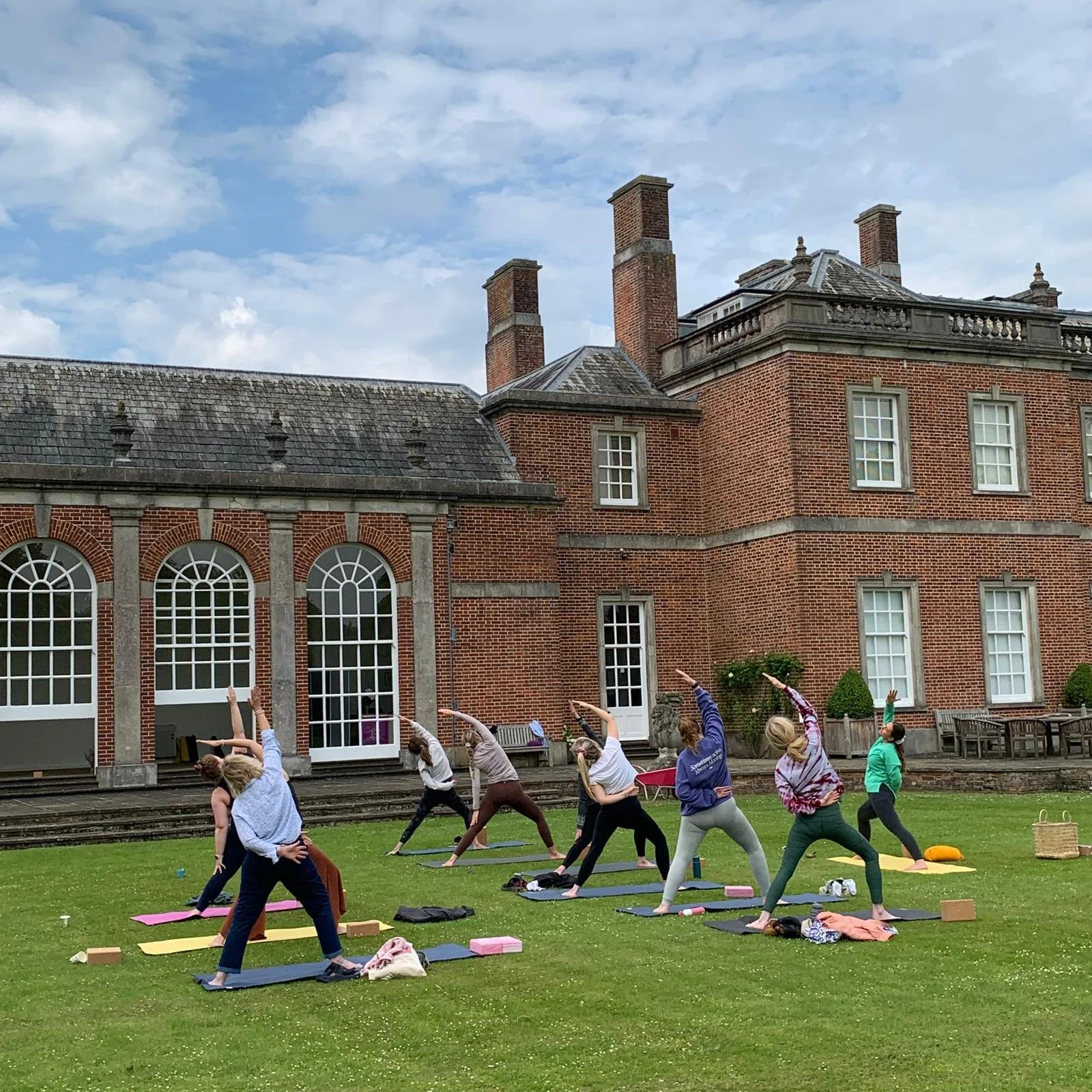 People practicing yoga outdoors on grass in front of a brick building with large windows, under a partly cloudy sky.