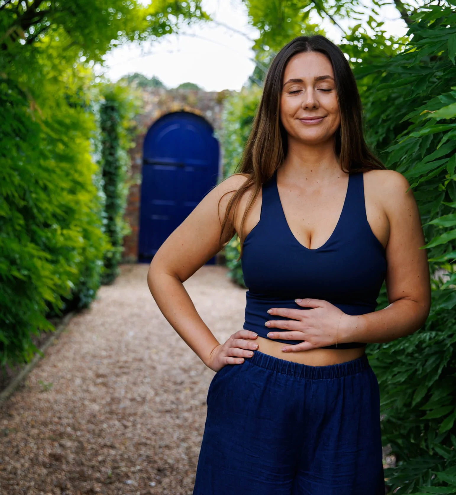 A young woman wearing a navy blue athletic top and matching navy blue pants, stands with her right hand on her hip and her left hand resting on her stomach, smiling with her eyes closed in a lush green garden pathway with a blue gate in the background.