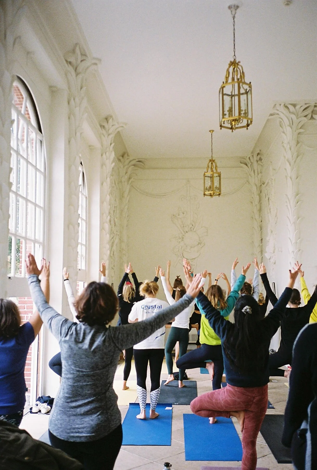 A group of people participate in a yoga or mindfulness class in a bright, spacious room with ornate white walls and large windows, overseen by a woman in pink guiding them.