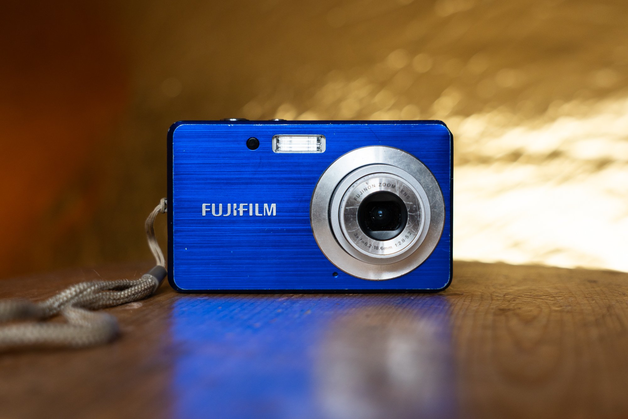 A bright blue digital Fujifilm camera sits on a wooden surface. In the background you can see a shiny gold reflector.