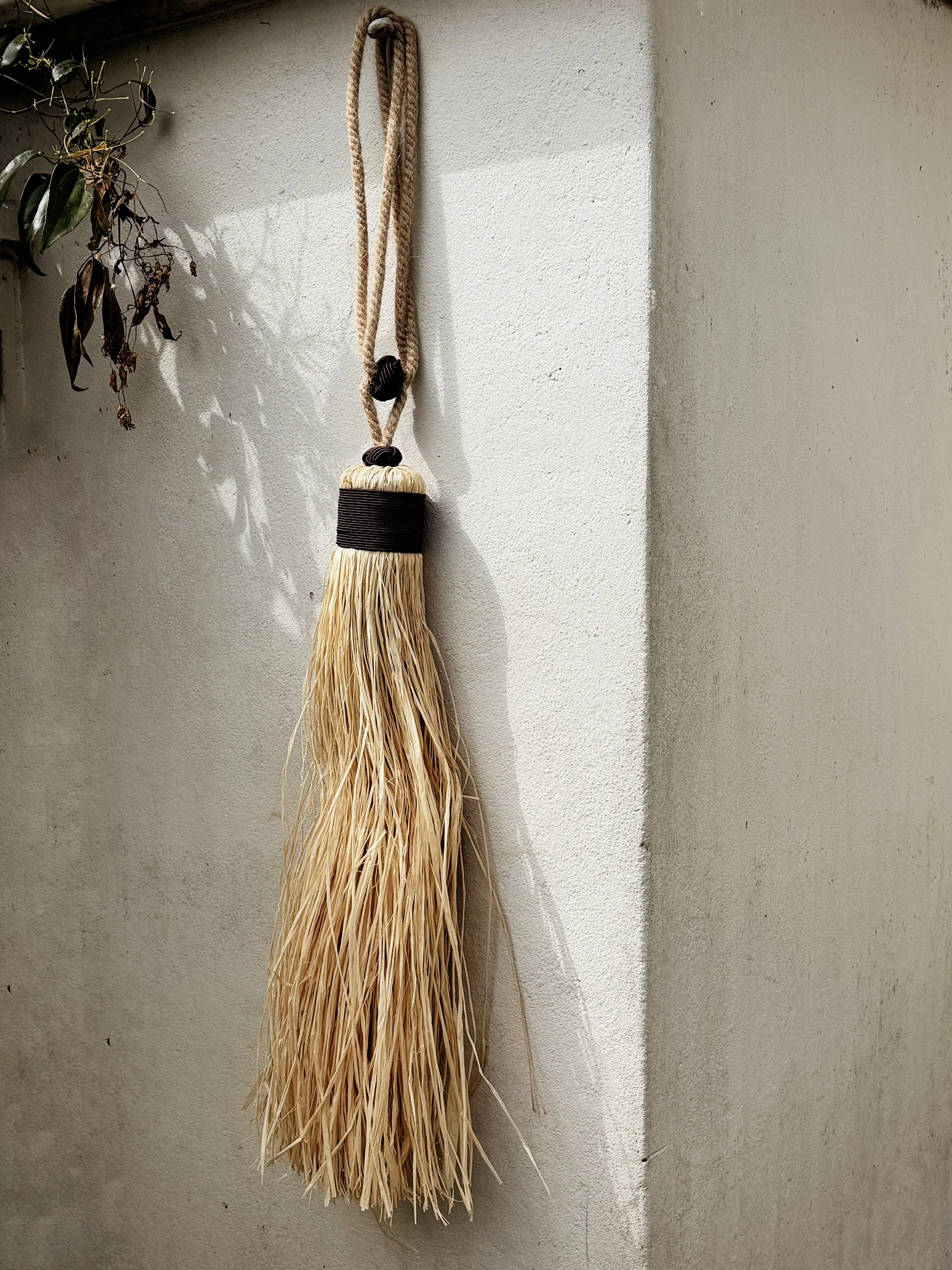 A straw broom with a black band hanging on a white wall near a corner, with dried leaves on a plant to the left.