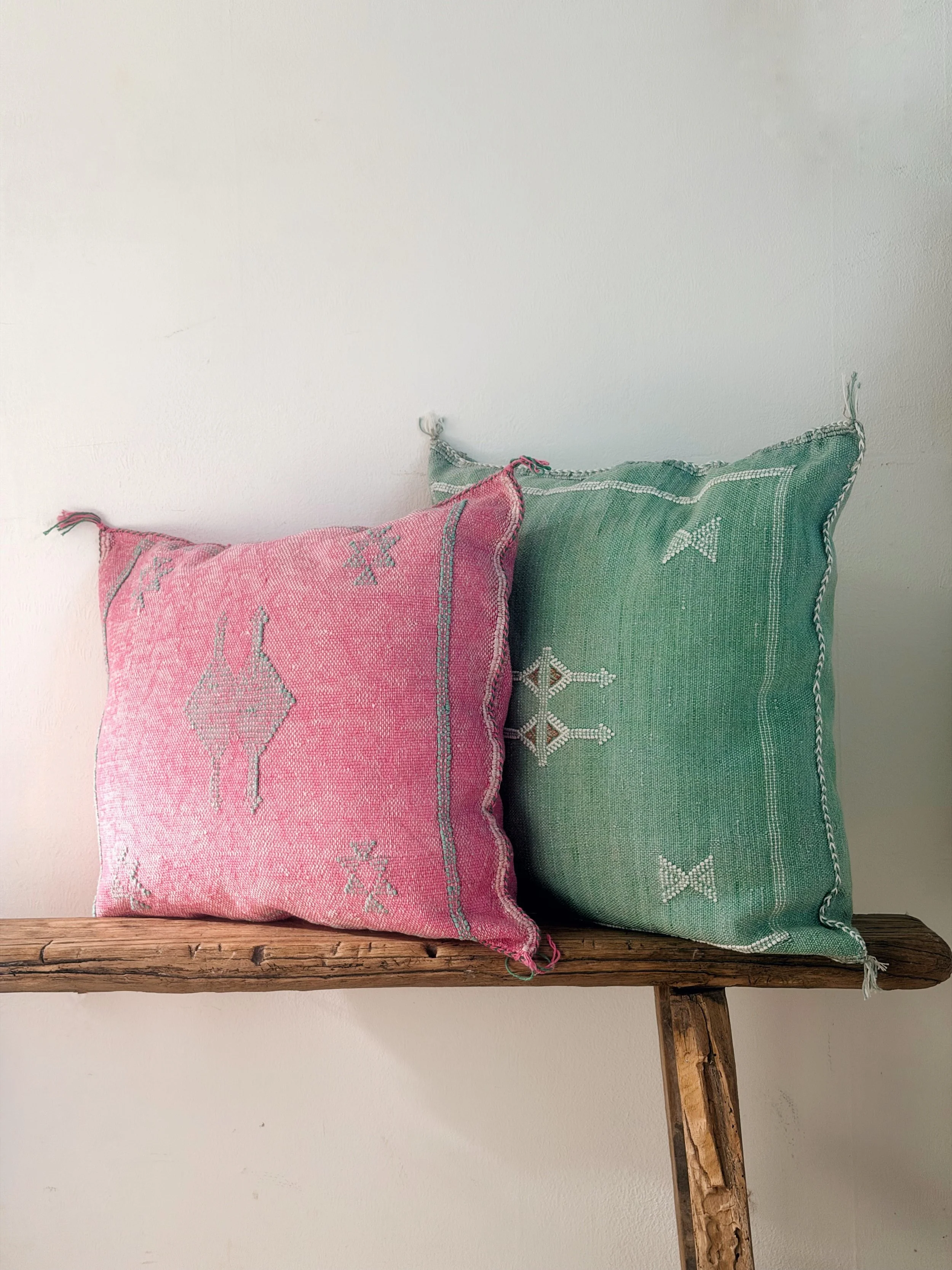 Two embroidered cushions, one pink and one green, placed on a rustic wooden shelf against a plain white wall.