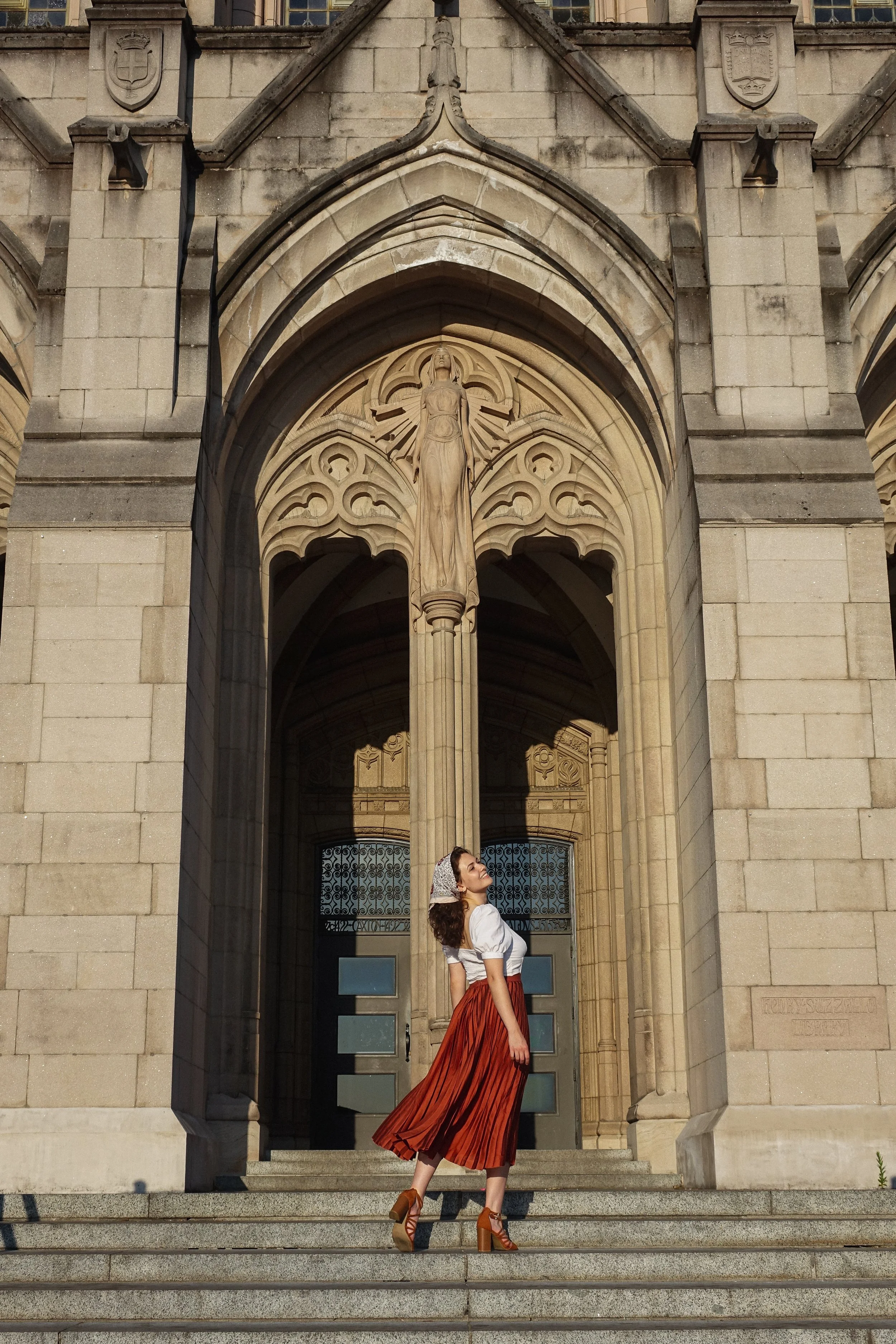 A woman in a long, orange skirt and high heels standing on stone steps in front of a historic building with gothic architecture, a large arched doorway, and a statue of a woman holding a cross above the entrance.
