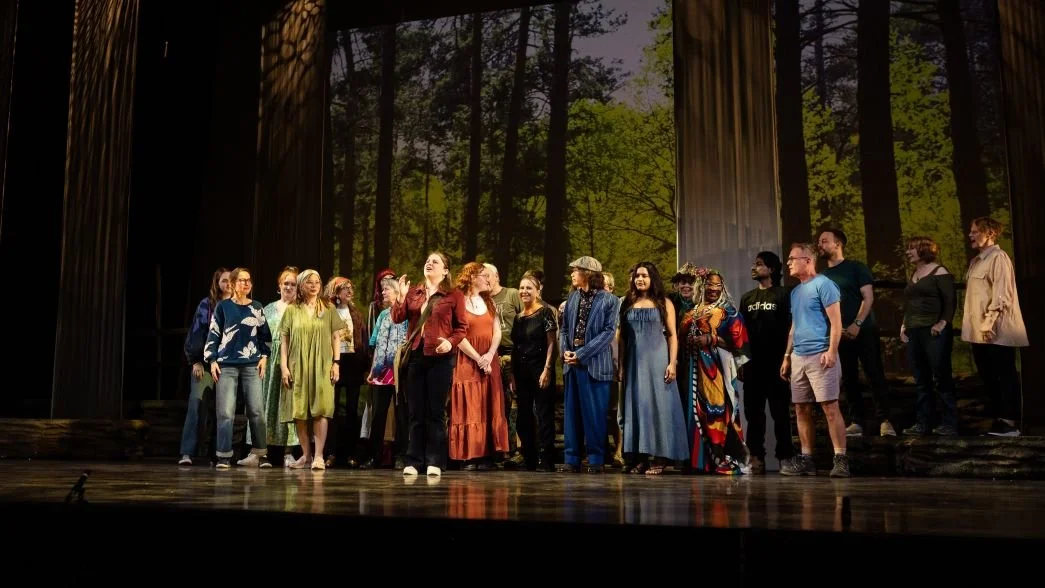 A group of diverse performers standing on a theater stage with a forest scene in the background.