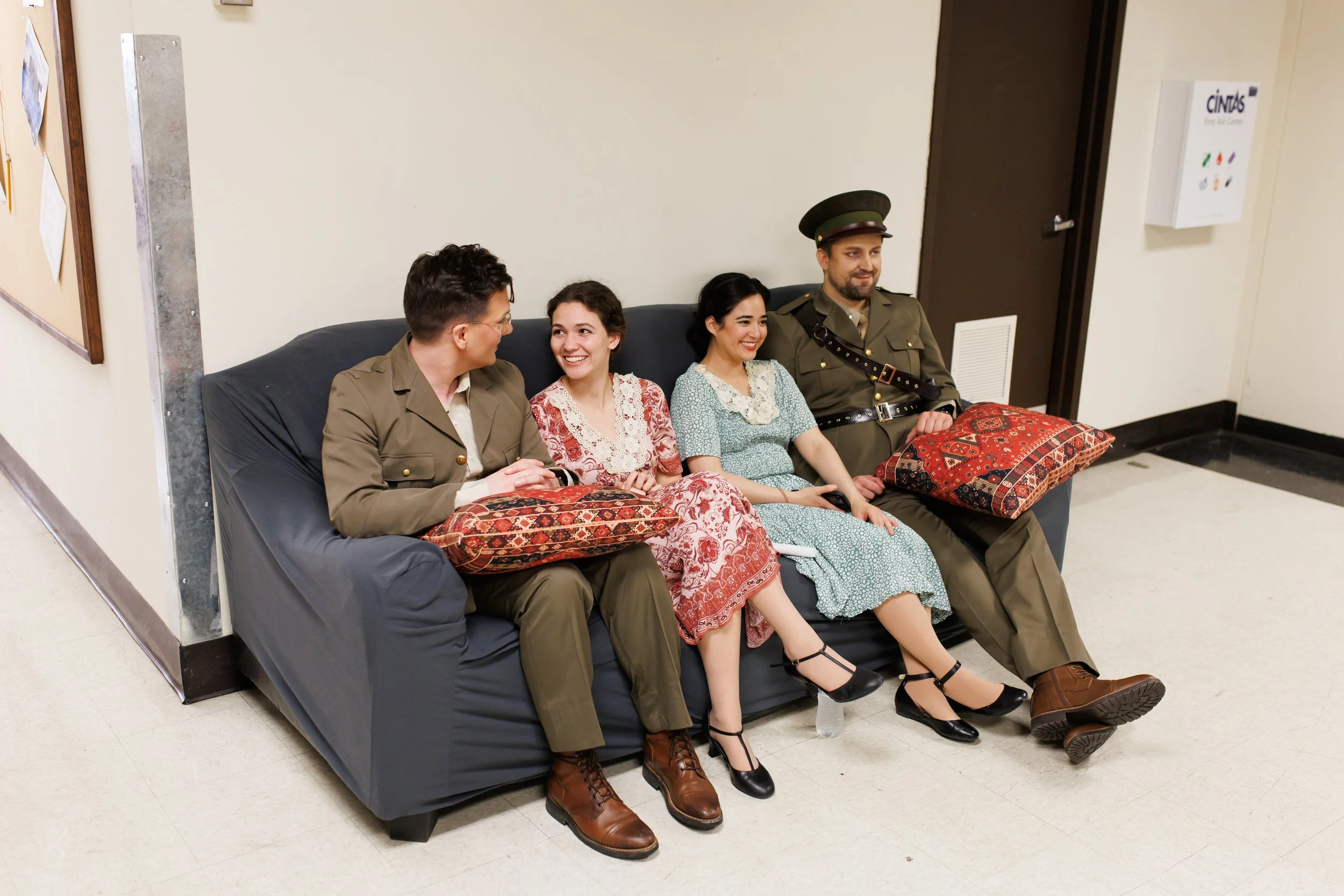 Four people sitting on a black couch: two men in military uniforms, two women in vintage dresses; they are smiling and talking, with pillows and a water bottle on the floor nearby.