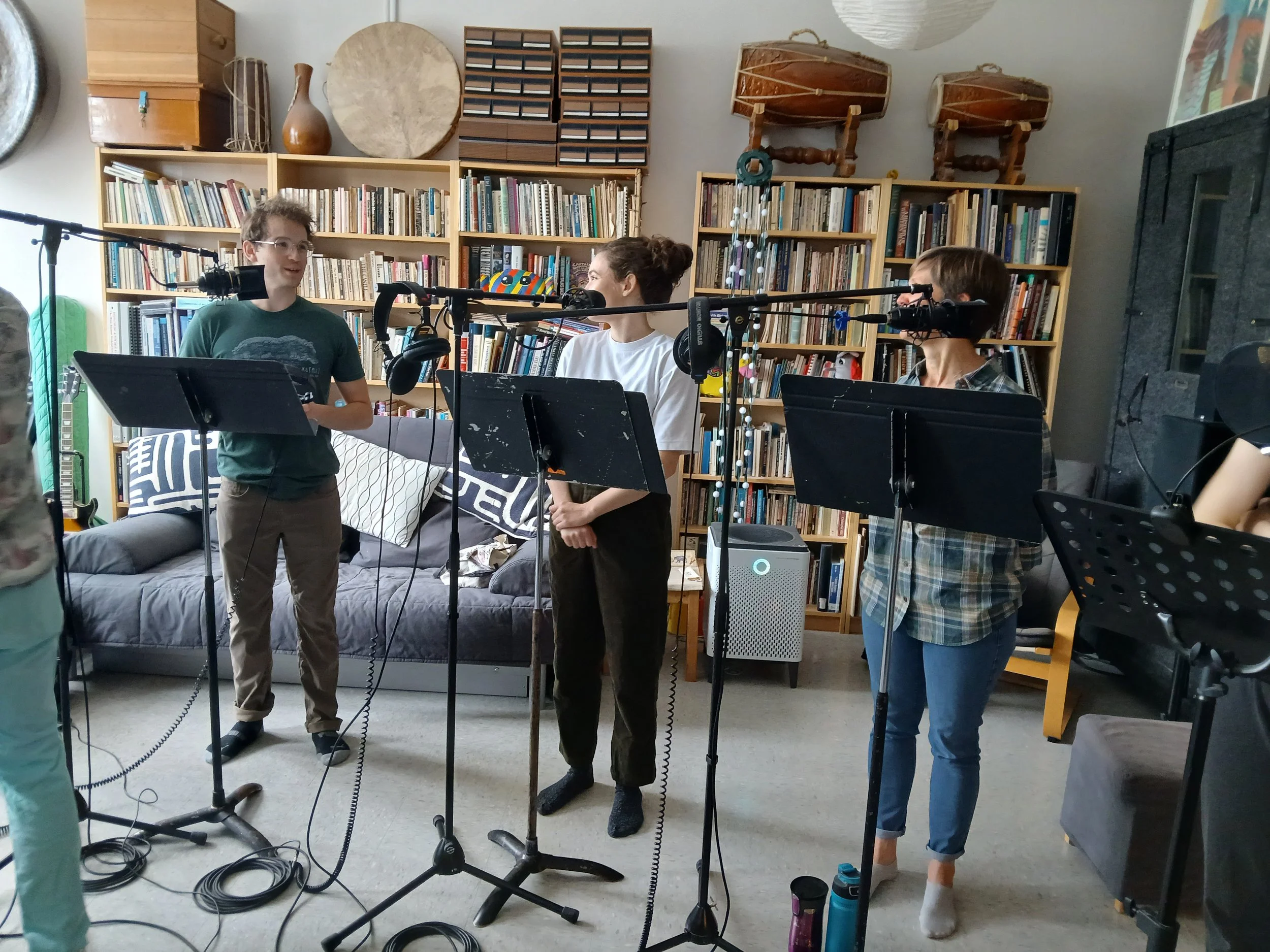 Three people are preparing to record or perform music in a cozy room with a bookshelf and musical instruments in the background. One person is speaking into a microphone, another is standing with arms crossed, and the third is wearing headphones near a microphone.