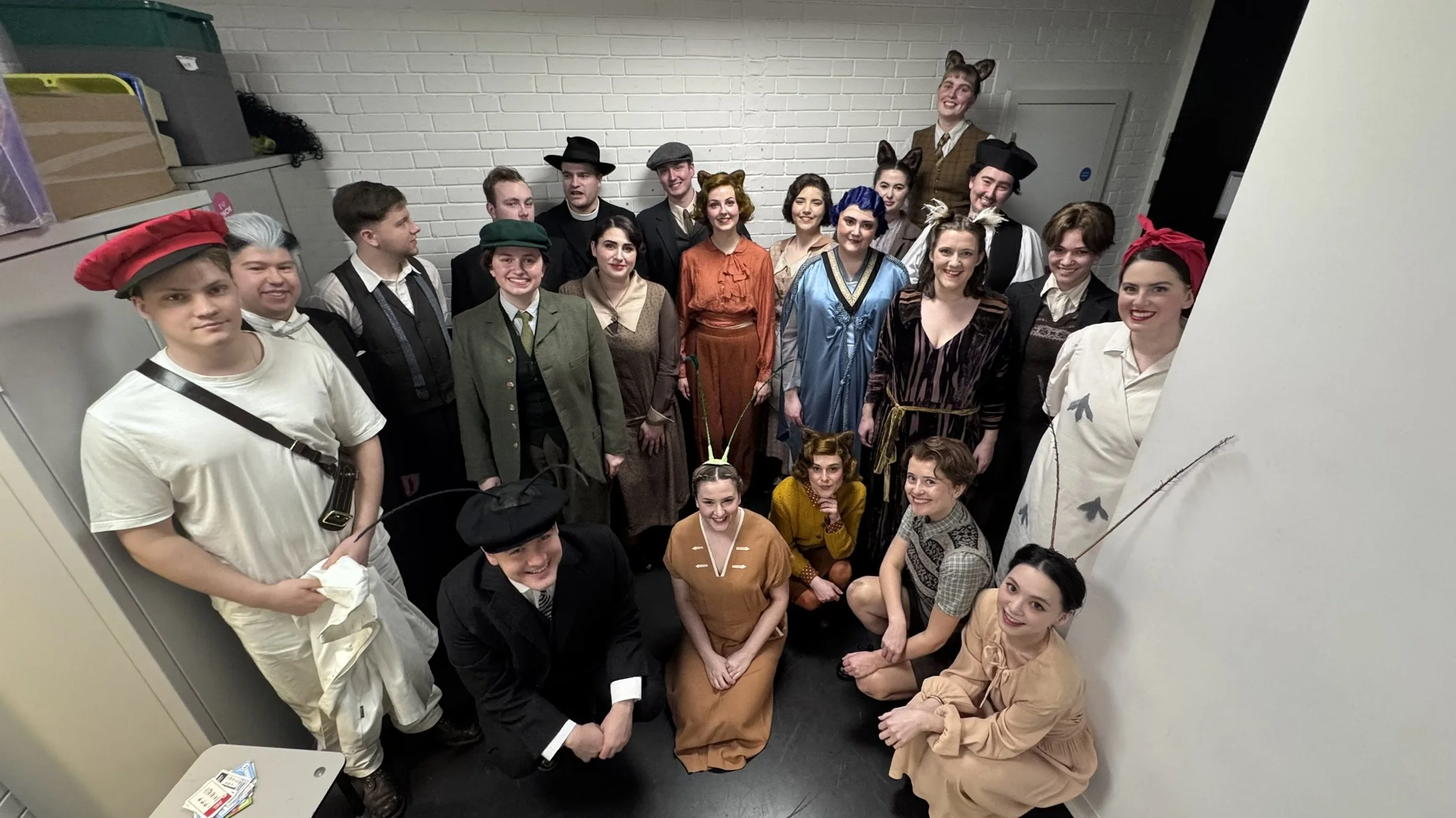 Group of people dressed in costumes, some with animal ears and tails, posing for a photo in an indoor setting with a white brick wall background.