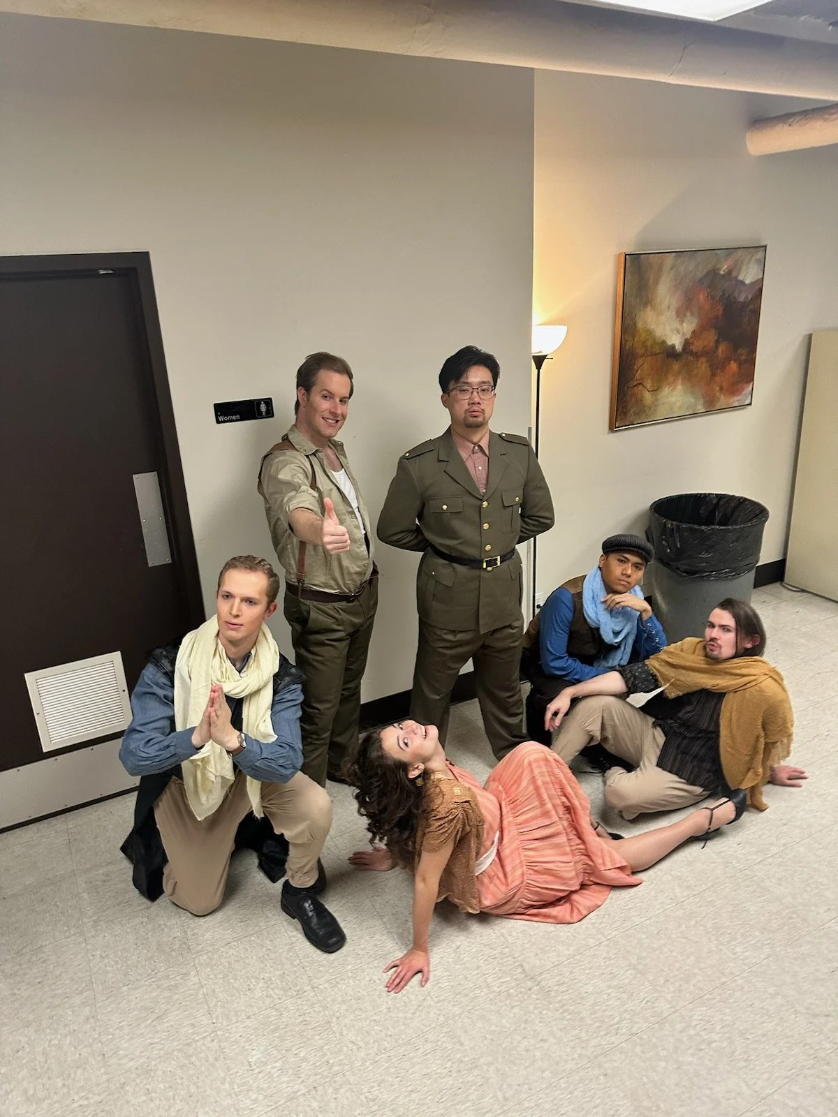 Group of six people dressed in various costumes posing in a hallway against a plain wall, with some sitting or lying on the floor, one giving a thumbs-up, and others with different facial expressions.