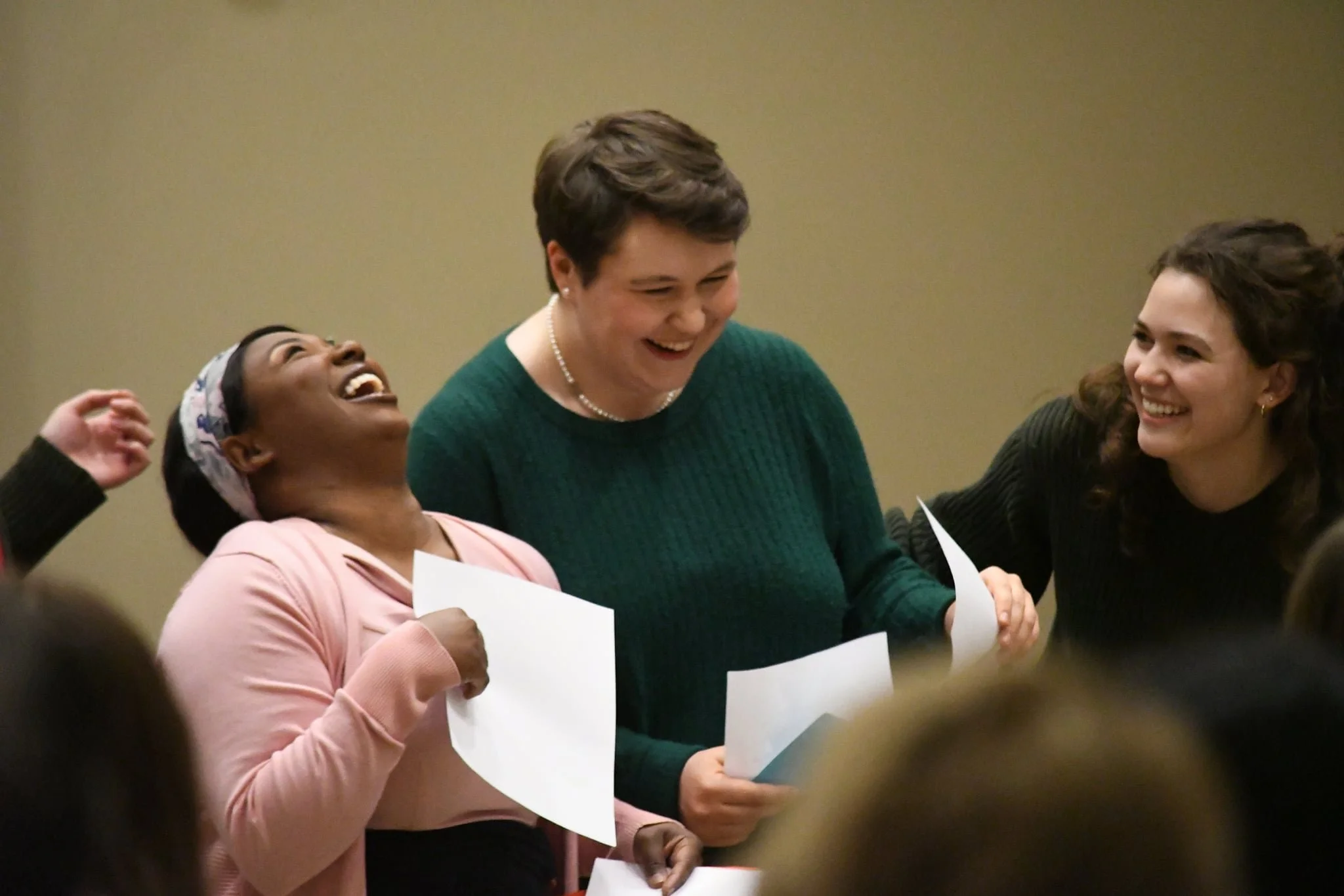 Three women are smiling and talking to each other while holding papers. They are dressed in business casual attire.