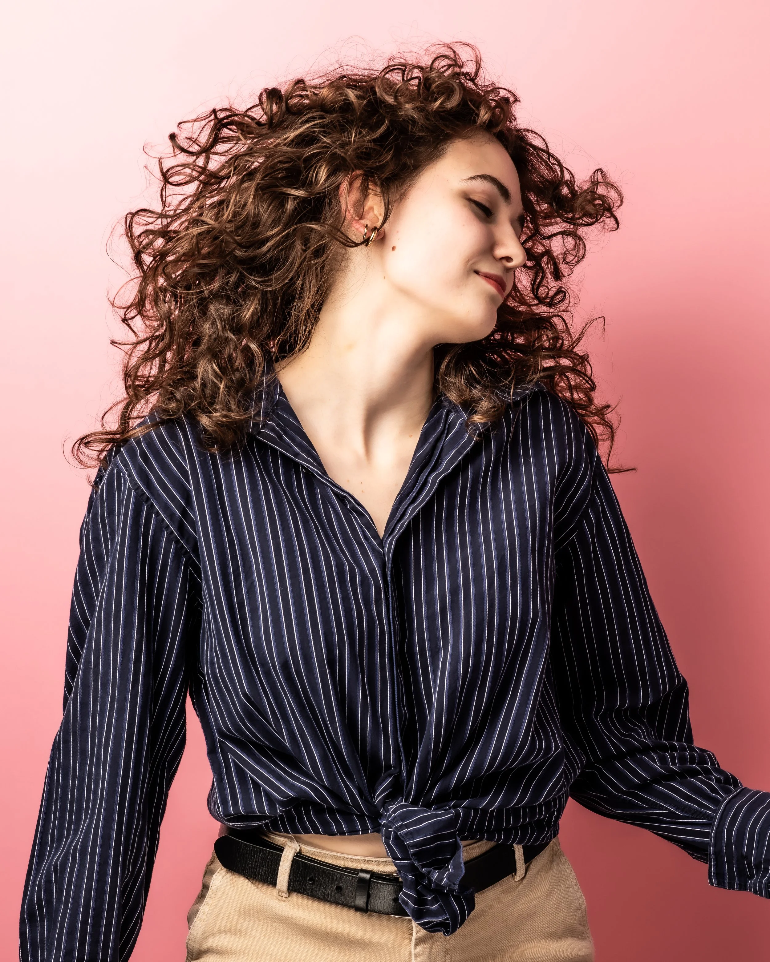 A young woman with curly hair, wearing a navy blue pinstripe shirt, beige pants, a black belt, and multiple earrings, posing against a pink background.