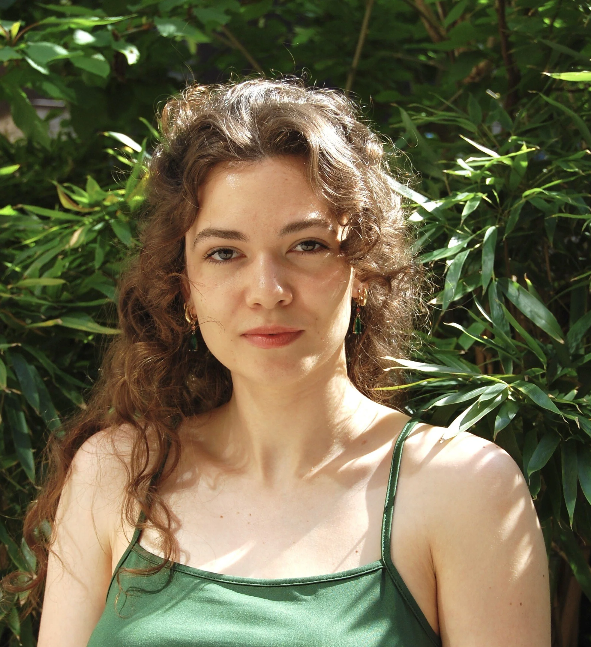 Close-up of a woman with curly hair wearing a green top, standing outdoors among green leafy plants.