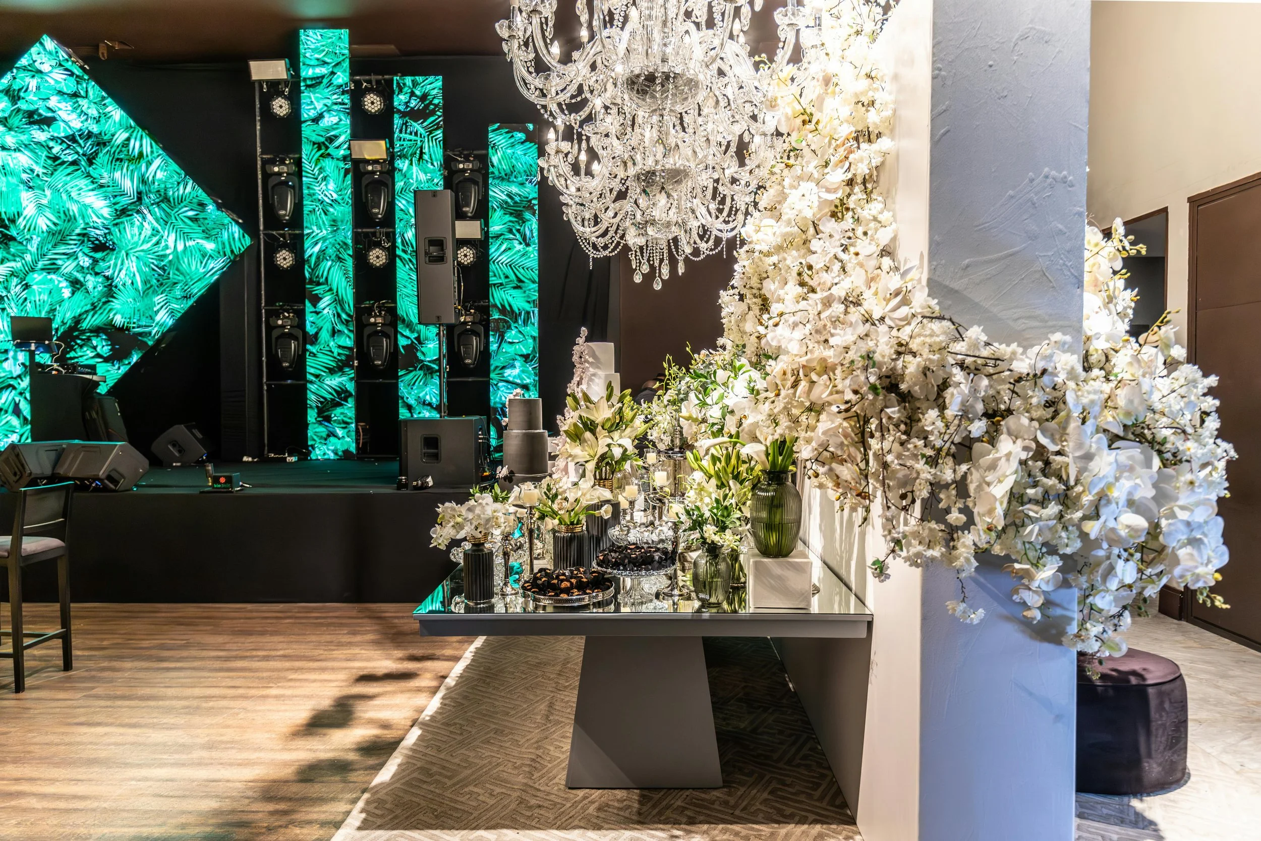 Modern event stage setup with illuminated green panels, crystal chandeliers, and lush white floral arrangements on a sleek display table.