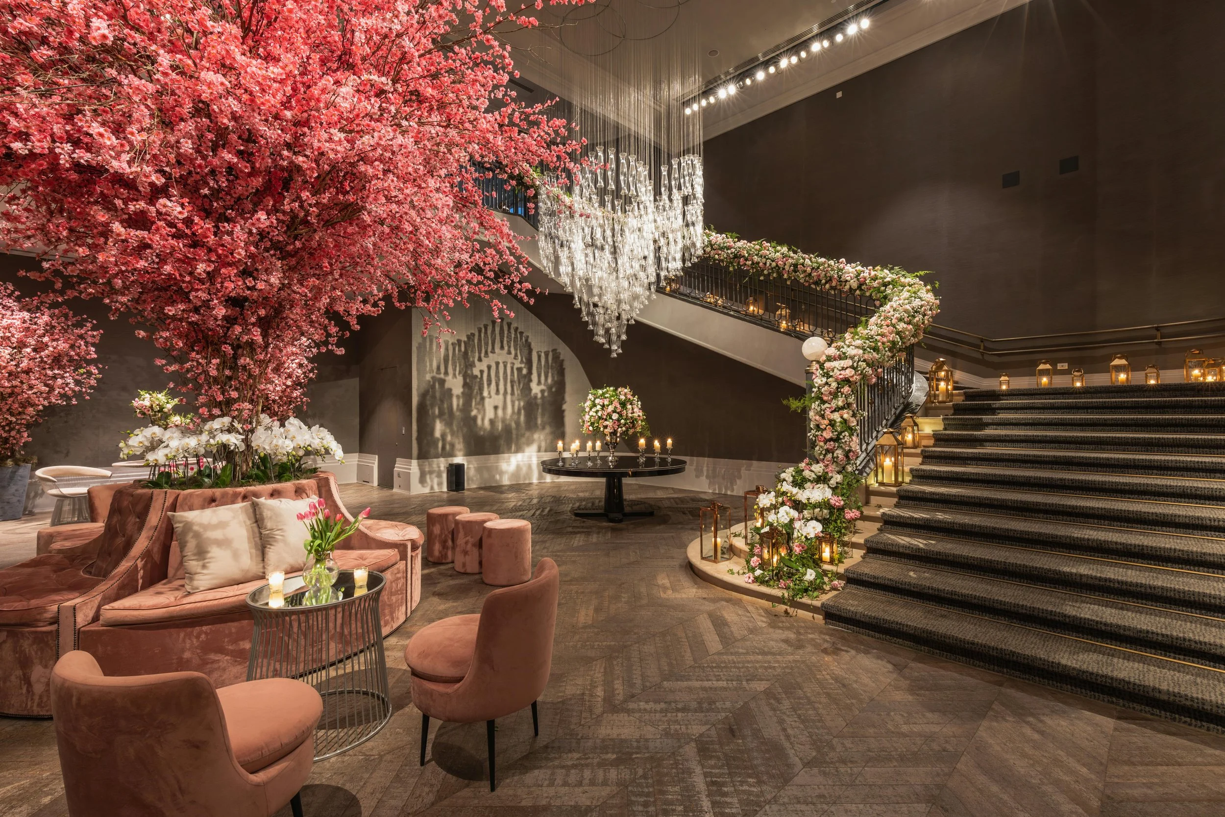Elegant event lounge with pink floral tree centerpiece, crystal chandeliers, and a grand staircase decorated with cascading flowers.
