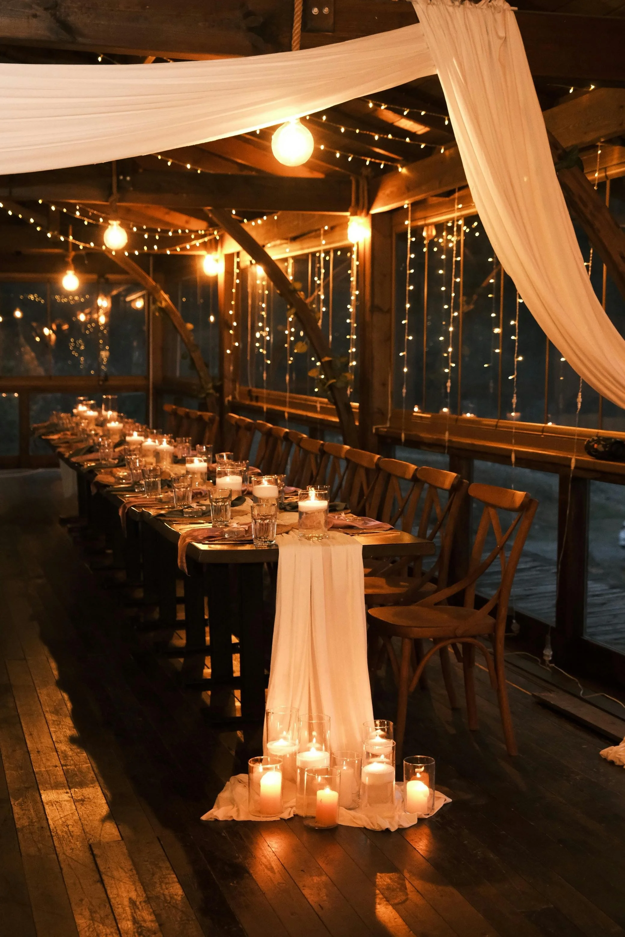 Elegant long banquet table set for an intimate evening event, featuring wooden chairs, soft candlelight in glass holders, sheer table runner, and warm string lights hanging along arched windows.