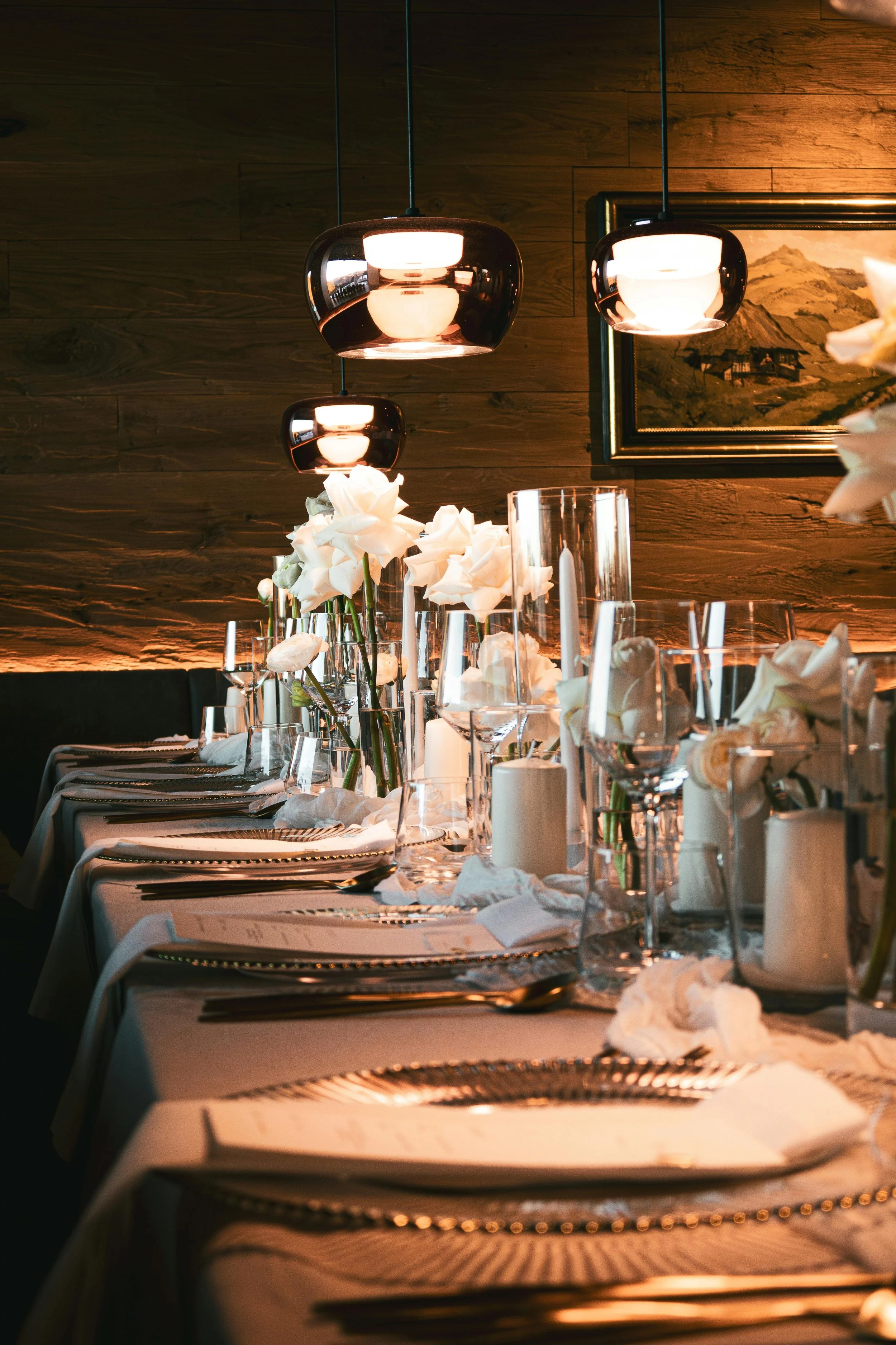 Intimate candlelit dining table with white floral arrangements, glass candle holders, and neatly arranged place settings creating a warm ambiance.