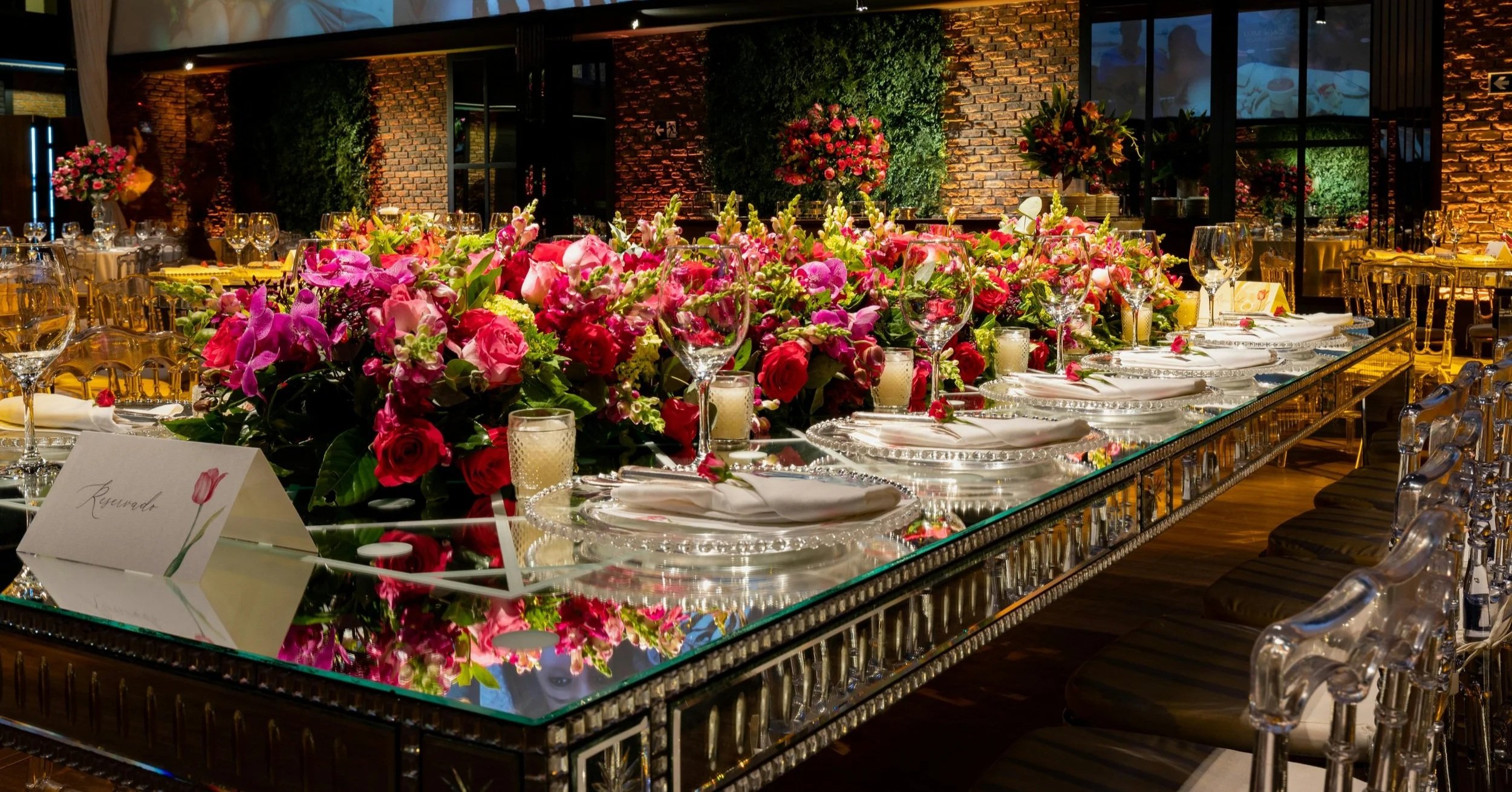 Luxurious banquet table with mirrored surface, vibrant pink and red floral centerpieces, and elegant place settings in an outdoor-style venue.