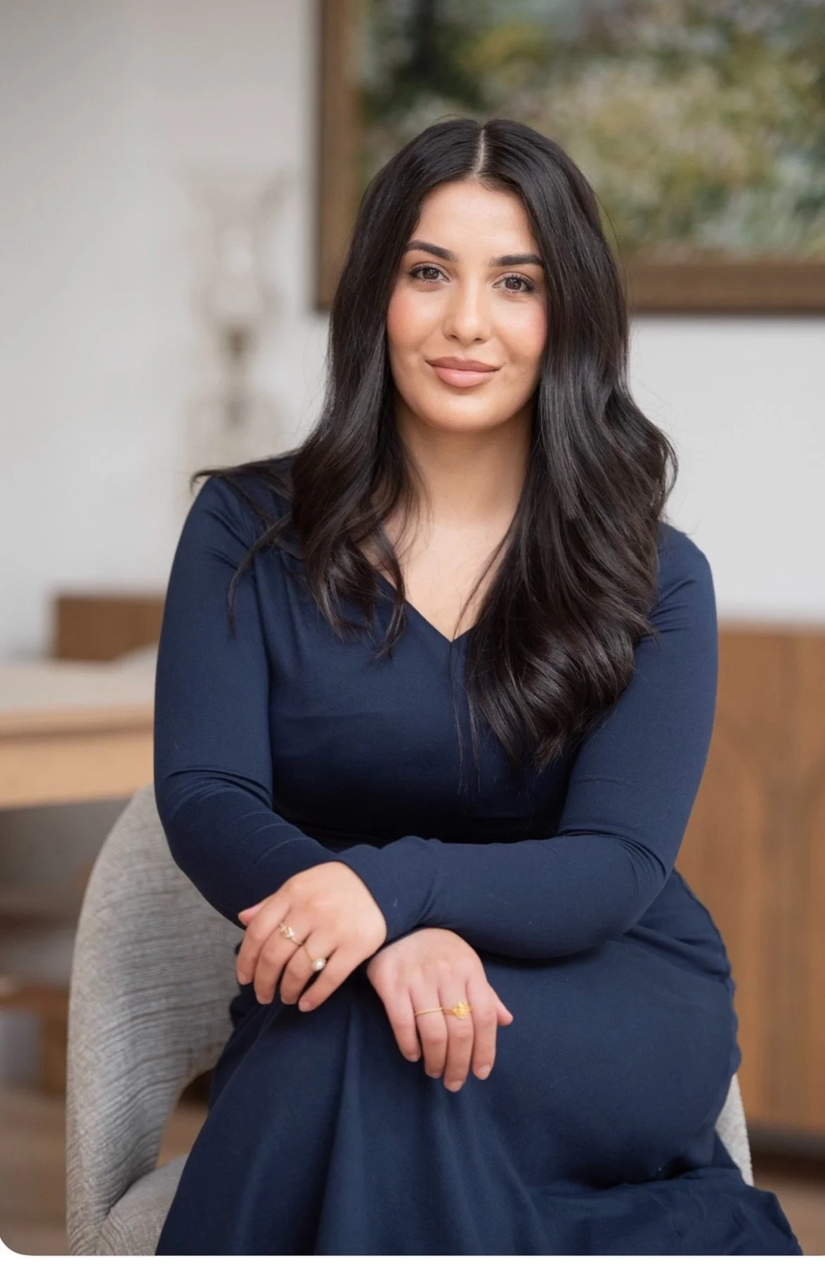 A woman with long dark hair, wearing a navy blue dress, seated on a chair indoors, smiling softly at the camera, with a background of a blurred wall and a window with greenery outside.