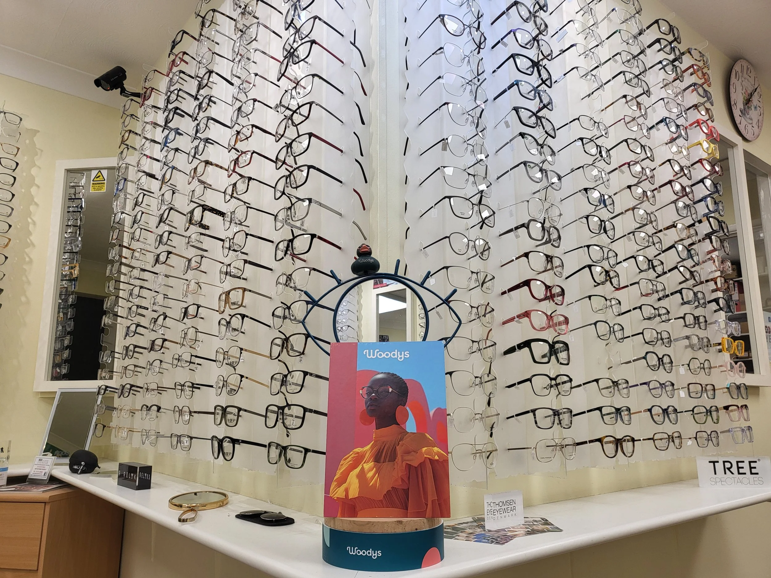 Display of eyeglasses on white wall in an eyewear store with a colorful promotional poster of a person wearing orange clothing and glasses.
