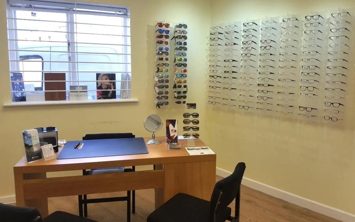 Eyeglasses display wall with multiple pairs of glasses and sunglasses in an optical shop, customer service desk with chairs, mirror, and window.