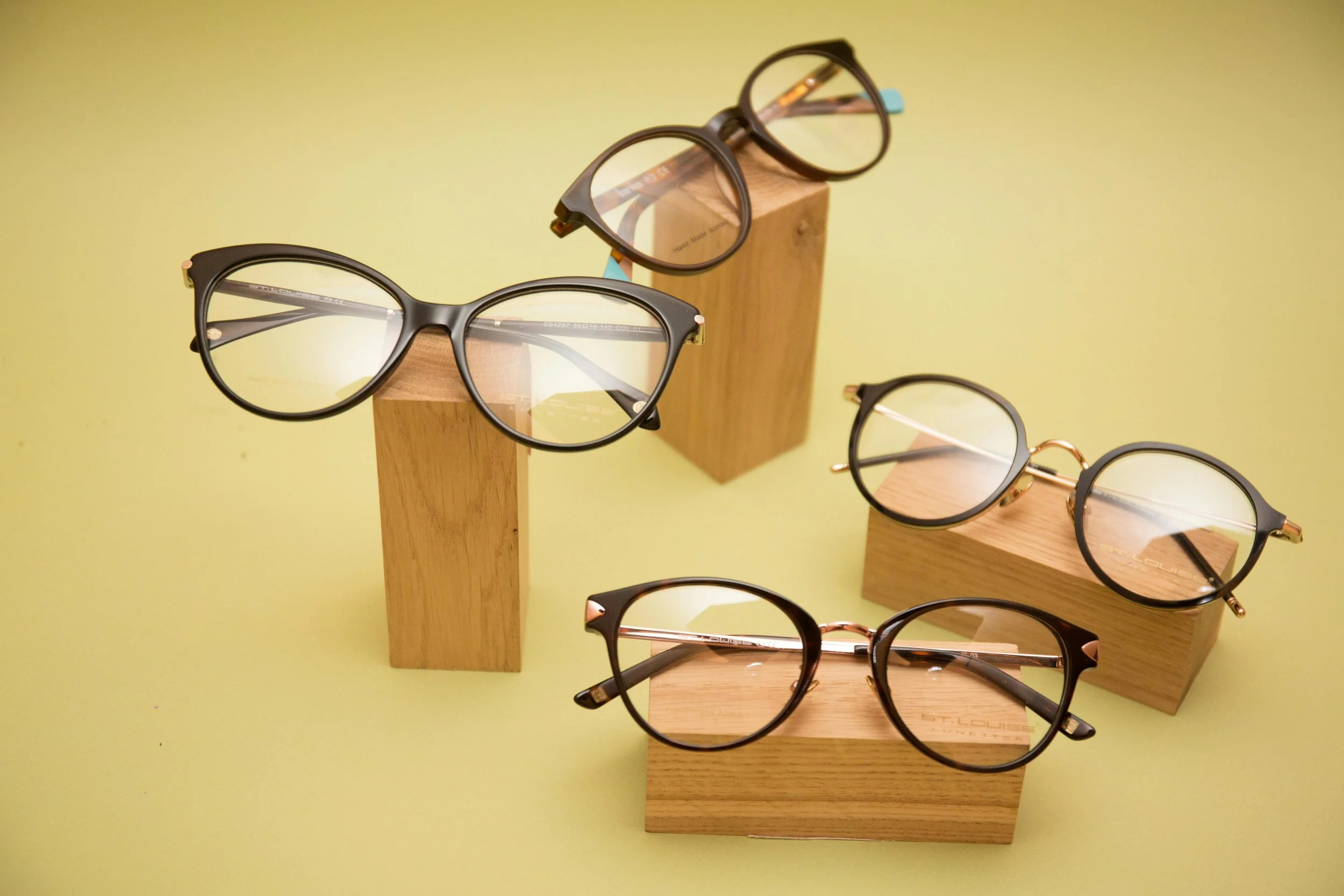 Multiple pairs of eyeglasses displayed on wooden stands against a yellow background.