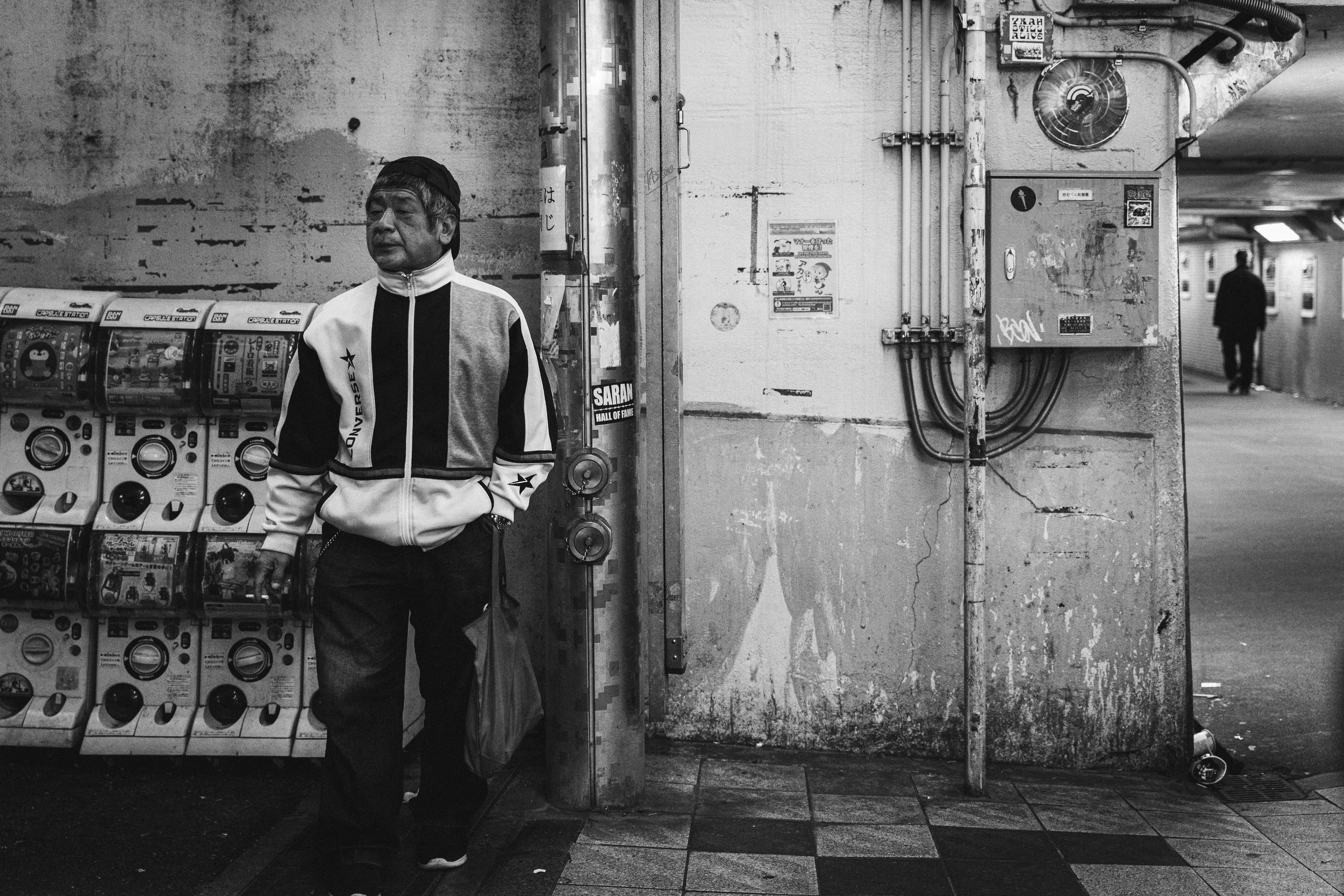 A black and white photo of a man standing in an underground subway station near a row of Japanese gashapon vending machines. The man is wearing a sporty jacket and a backward cap, looking contemplative. The background includes a wall with peeling pai