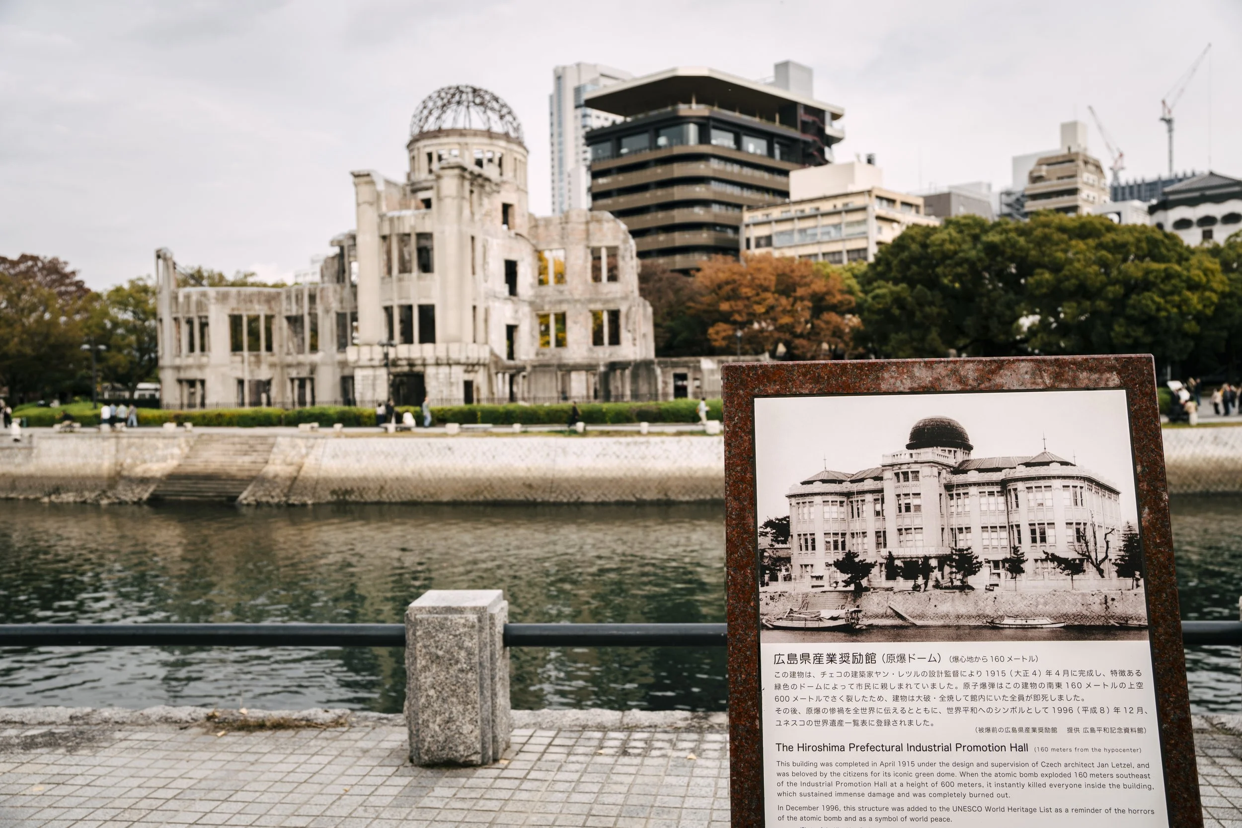 Black and white photograph of the Hiroshima Prefectural Industrial Promotion Hall, a historic building with a distinctive dome-shaped roof, displayed on an outdoor sign by a river with trees in fall foliage. Modern buildings and partly cloudy sky are