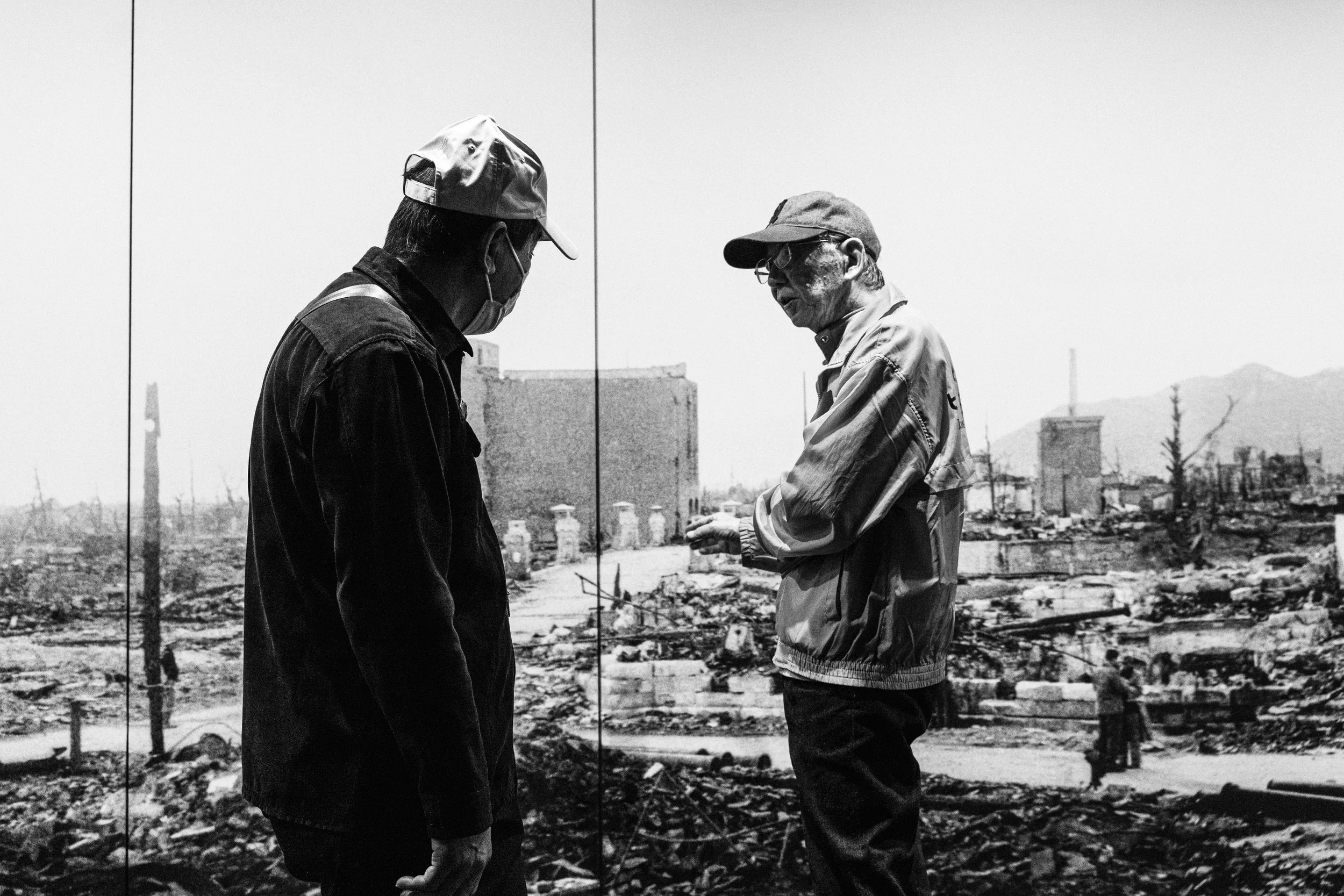 Two men with hats and glasses engage in conversation amidst the ruins of a destroyed area, with debris and damaged buildings in the background.