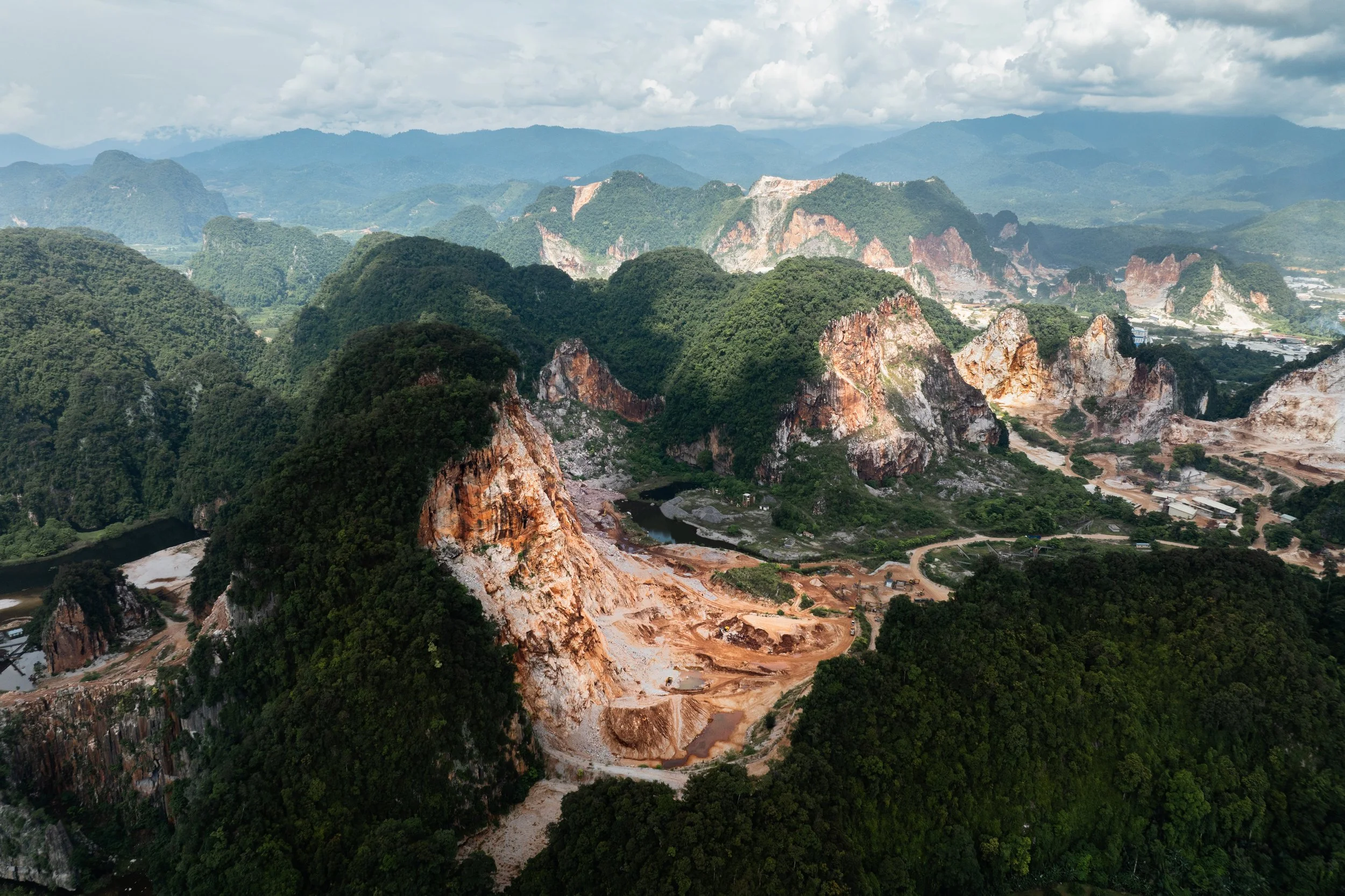 Perak, Malaysia — Ancient limestone karst peaks, stripped on one side and forested on the other. Quarry roads thread through the valley below. The mountain being taken apart, piece by piece, seen from above.