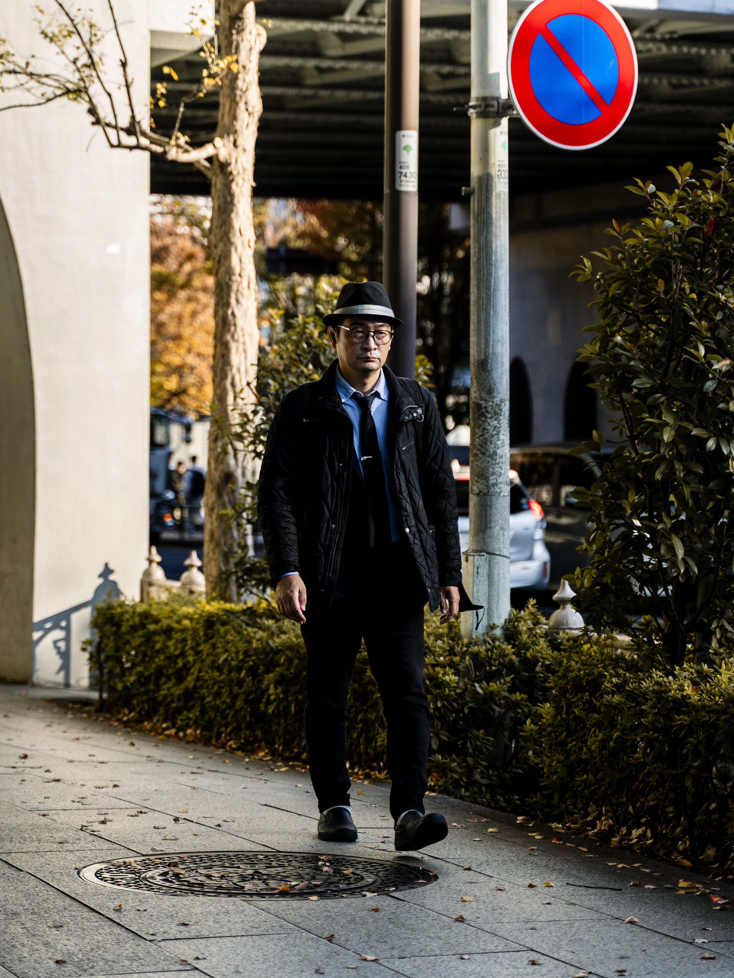 A man walking on a sidewalk, dressed in black clothing, wearing glasses and a fedora hat, with autumn trees and parked cars in the background, and a no-parking sign on a pole.