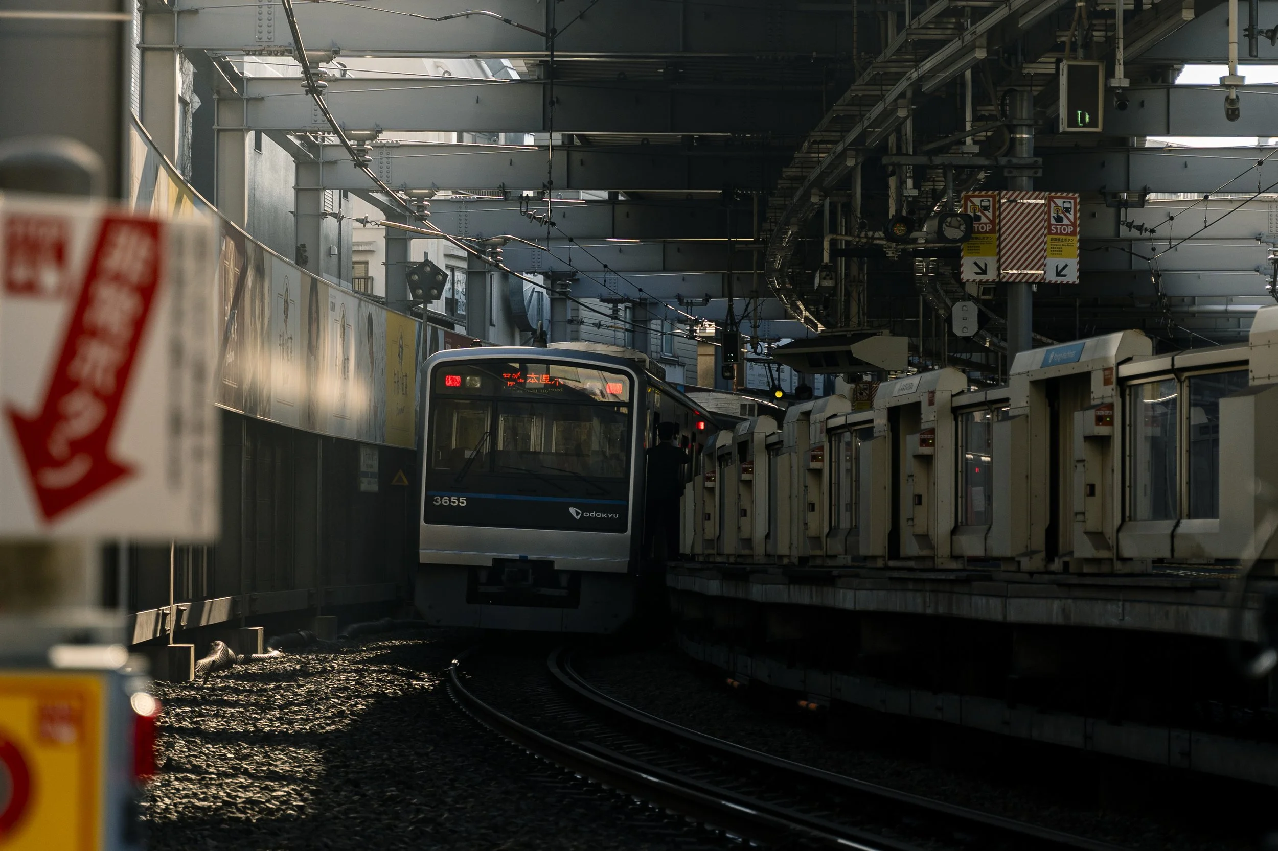 Odakyu 3000 Series, Yoyogi Hachiman, Tokyo — The commuter train rounds the curve into the station, headlights cutting through the shadows. Platform doors, emergency signs, late afternoon light. The unglamorous machinery of a city that never stops.