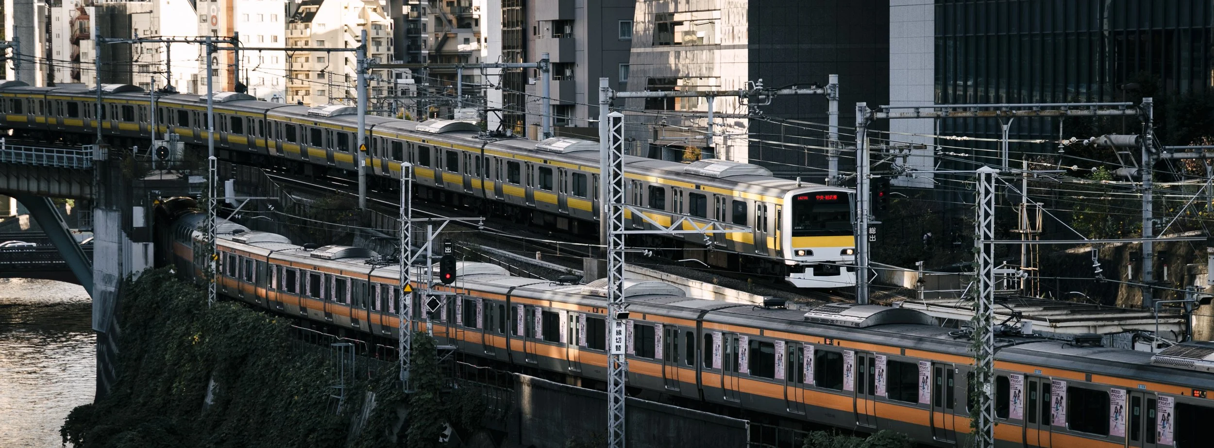 Cityscape with two trains traveling on separate elevated tracks over a river, surrounded by tall modern buildings and urban infrastructure.