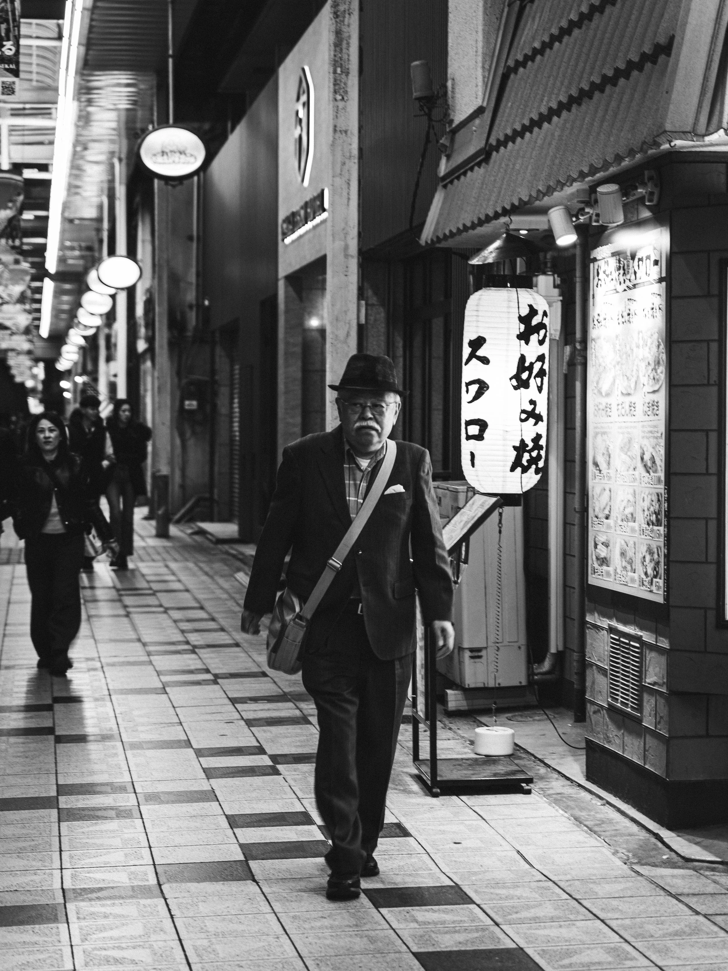 A man with glasses and a hat walking down a tiled sidewalk in an urban shopping district at night, with illuminated signs and other pedestrians in the background.