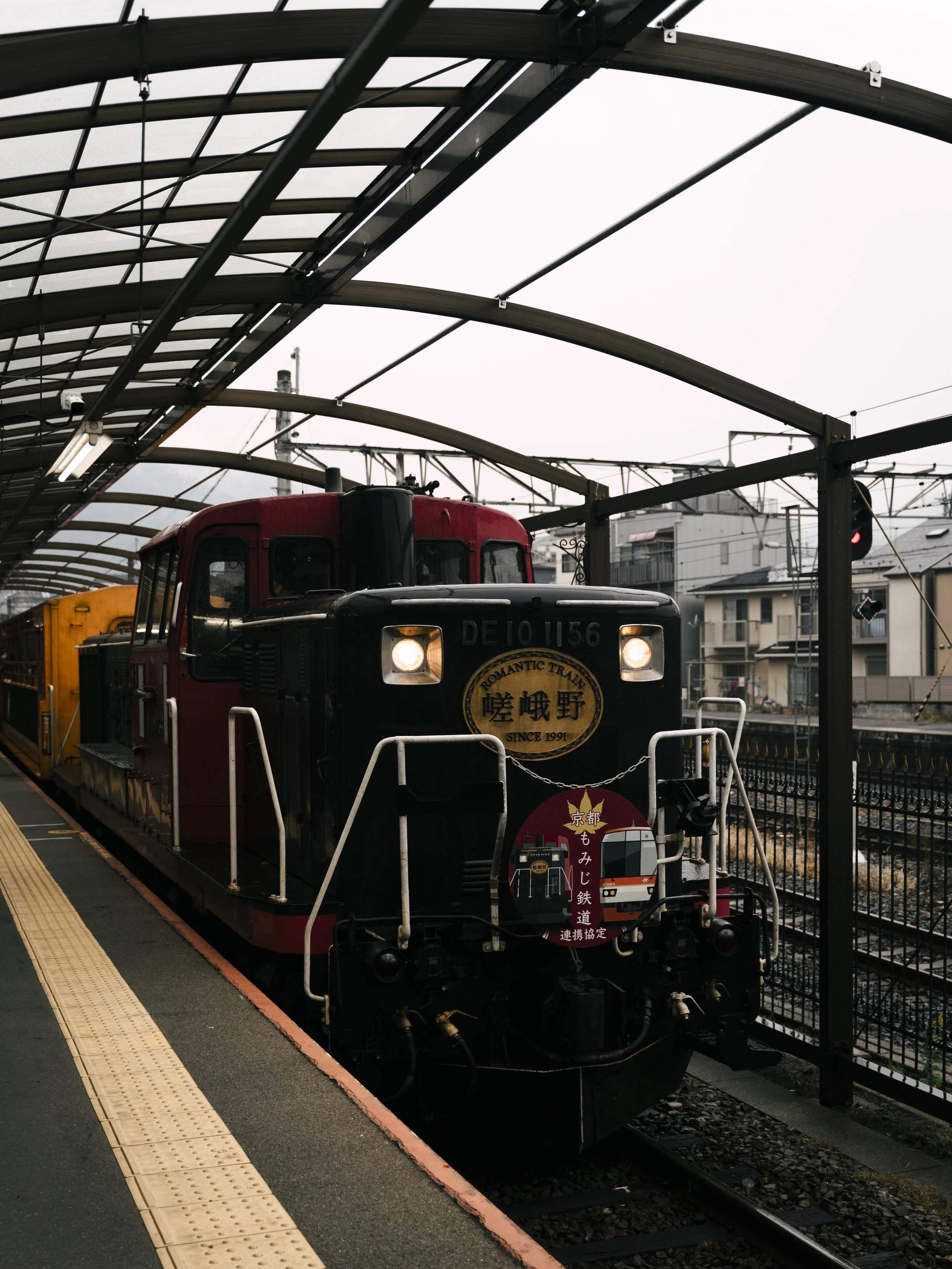 Sagano Romantic Train, Arashiyama, Kyoto — The DE10 1156 diesel locomotive waits beneath the arched station canopy, headlights glowing in the grey morning. Running since 1991 through the Hozugawa gorge — a journey where the train is entirely the poin