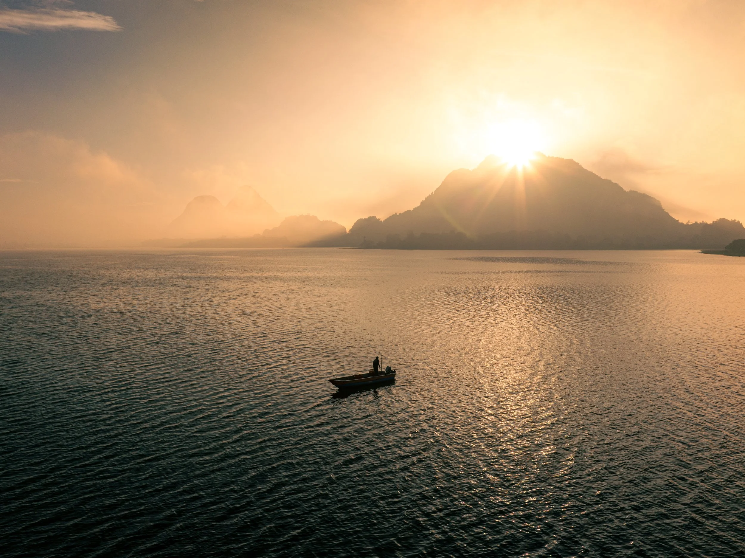 A person in a small boat on a calm body of water at sunset or sunrise, with the sun partially hidden behind a mountainous landscape in the background.