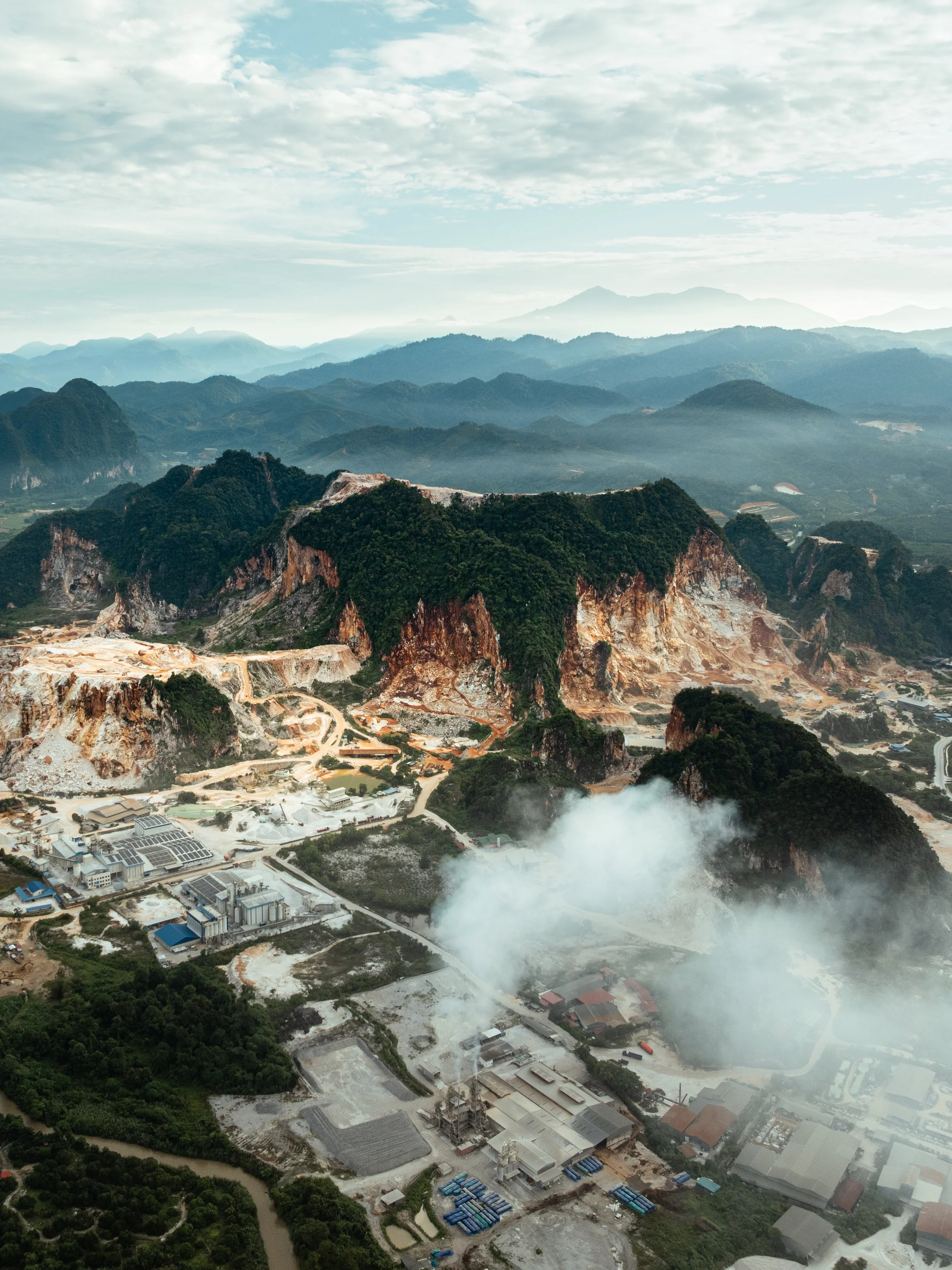 Industrial smoke rises from processing facilities nestled among quarried limestone hills in Simpang Pulai, Perak, with distant mountain ranges in the background.