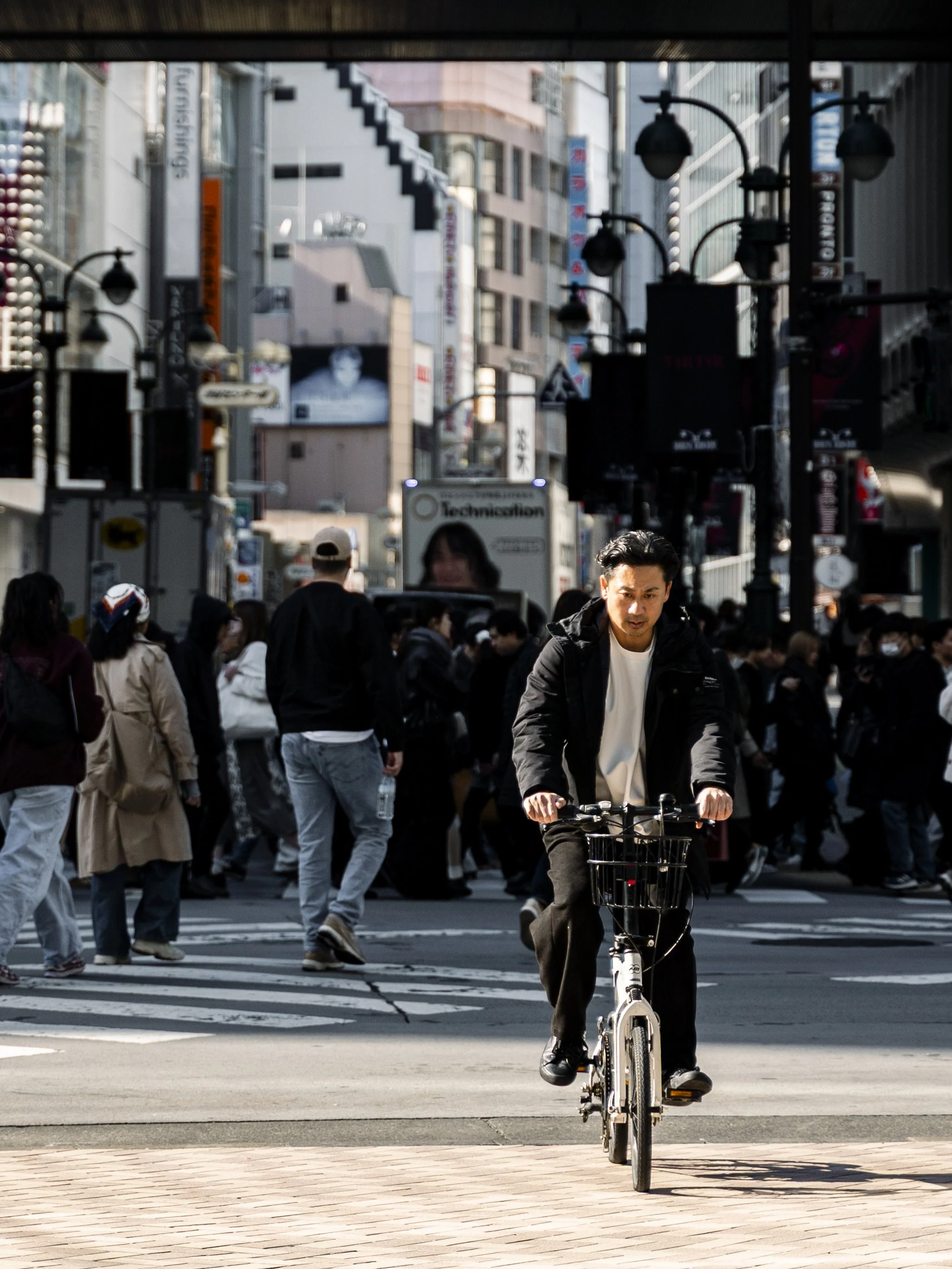 A man riding a bicycle on a city street with a crowd of people crossing the intersection, skyscrapers, and streetlights in the background.