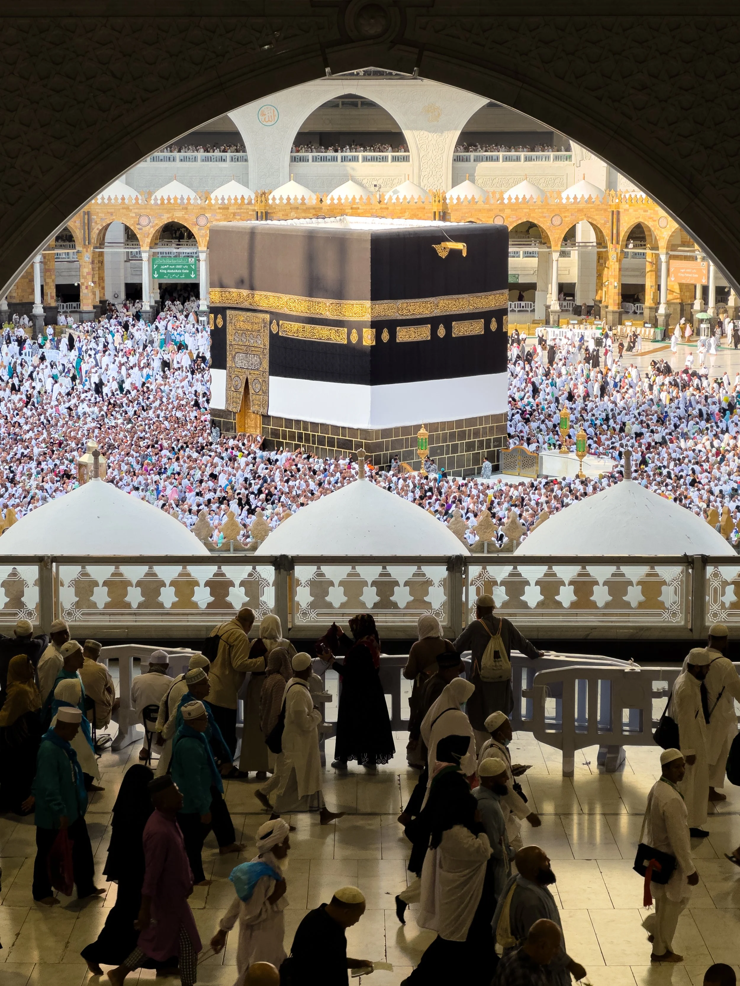 Masjidil Haram, Mecca, Saudi Arabia — The Kaabah fills the archway, draped in the Kiswah, surrounded by thousands. Silhouettes watch in stillness from the foreground. The scale only becomes real when you stop and look properly.
