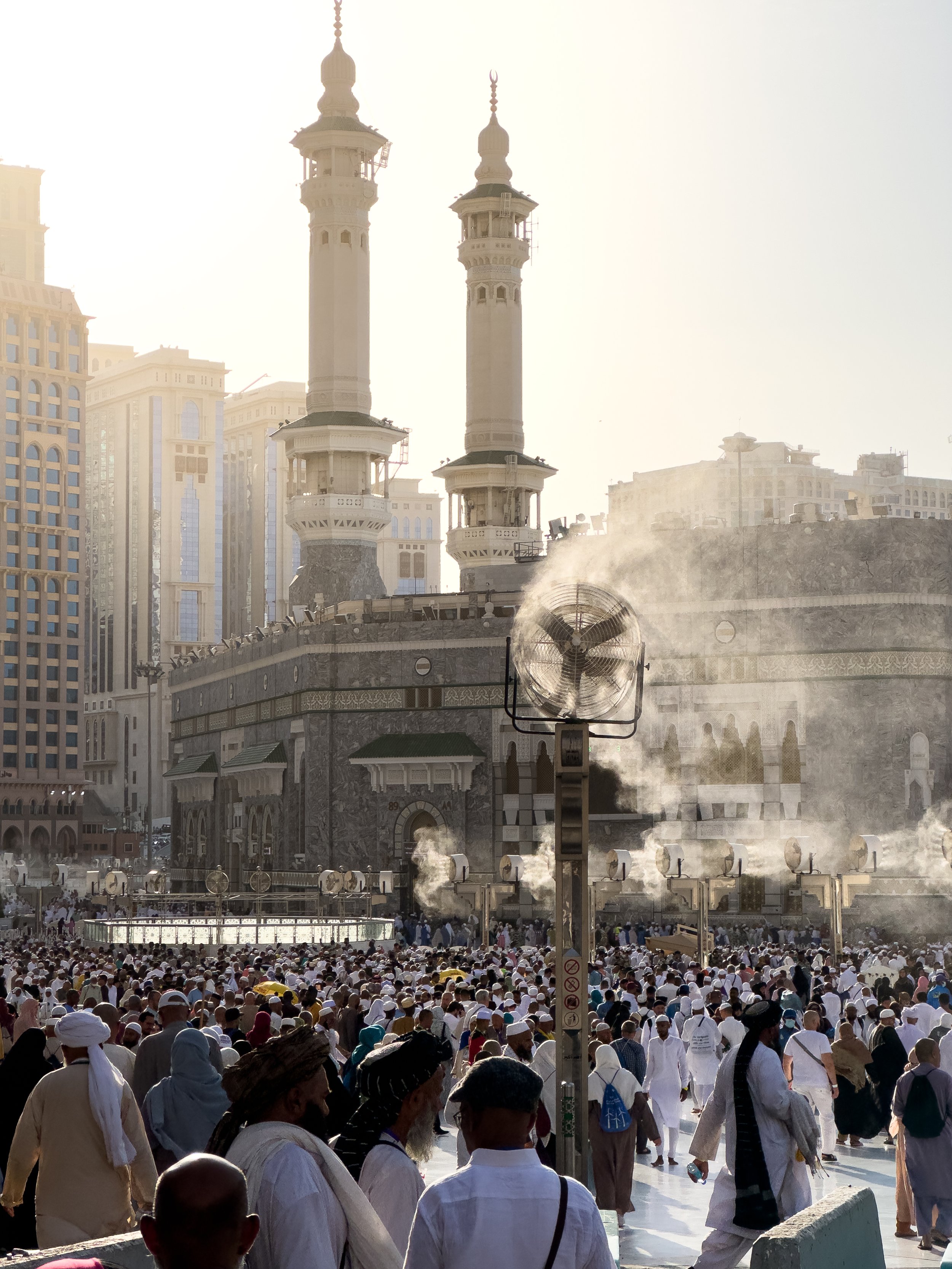 Masjidil Haram, Mecca, Saudi Arabia — Mist fans spray cooling water across the outer courtyard as thousands of pilgrims move in the midday heat. The minarets rise through the haze behind. Endurance, made slightly more bearable.