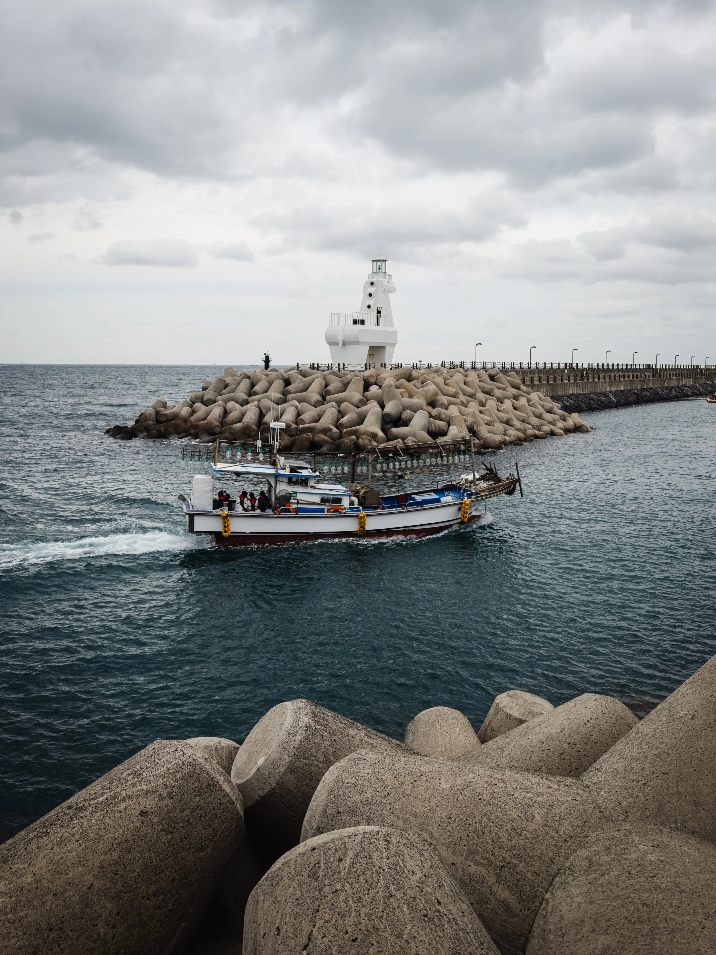 Jeju Island, South Korea — A squid fishing boat heads out past the lighthouse and the tetrapods. The sea is grey, the sky is grey, the boat is going anyway.